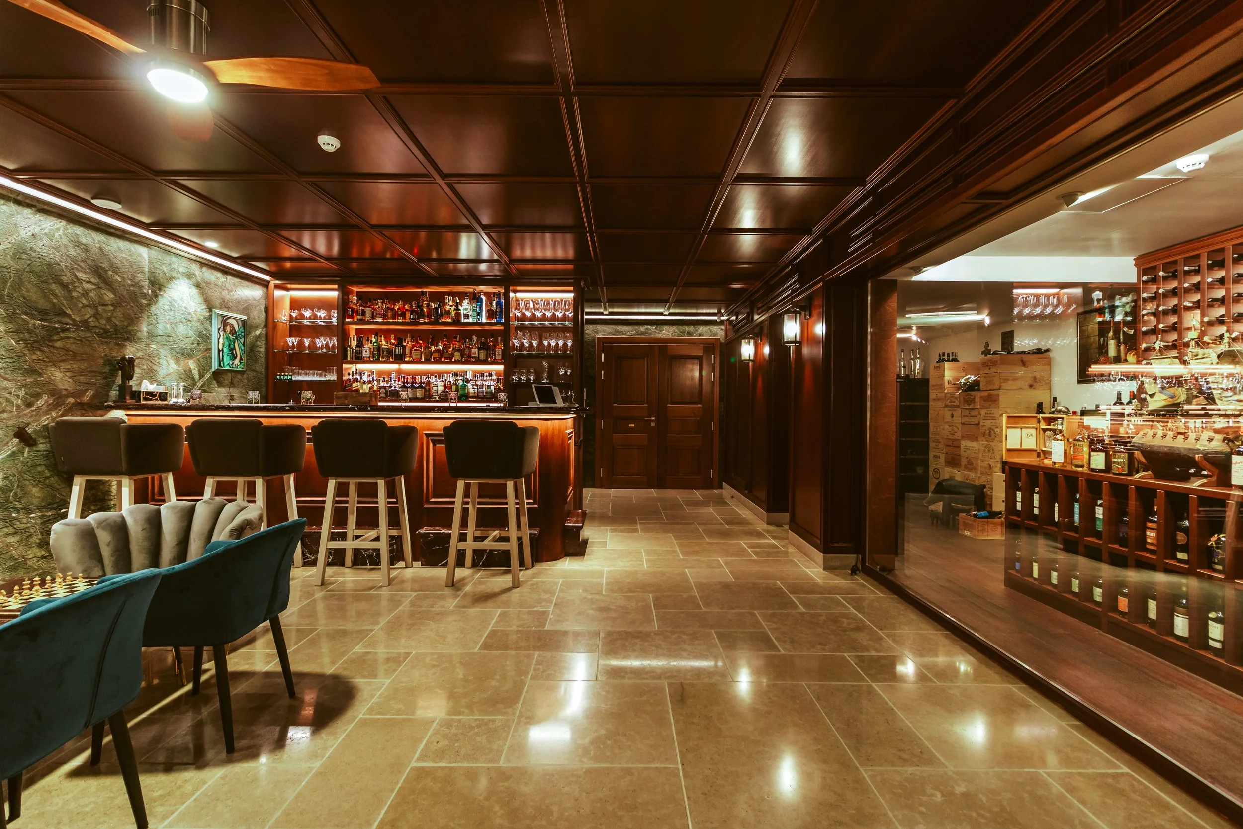 Interior of a bar with a wooden counter, bar stools, glass shelves with liquor bottles, and a cozy seating area with chairs and a chess set.