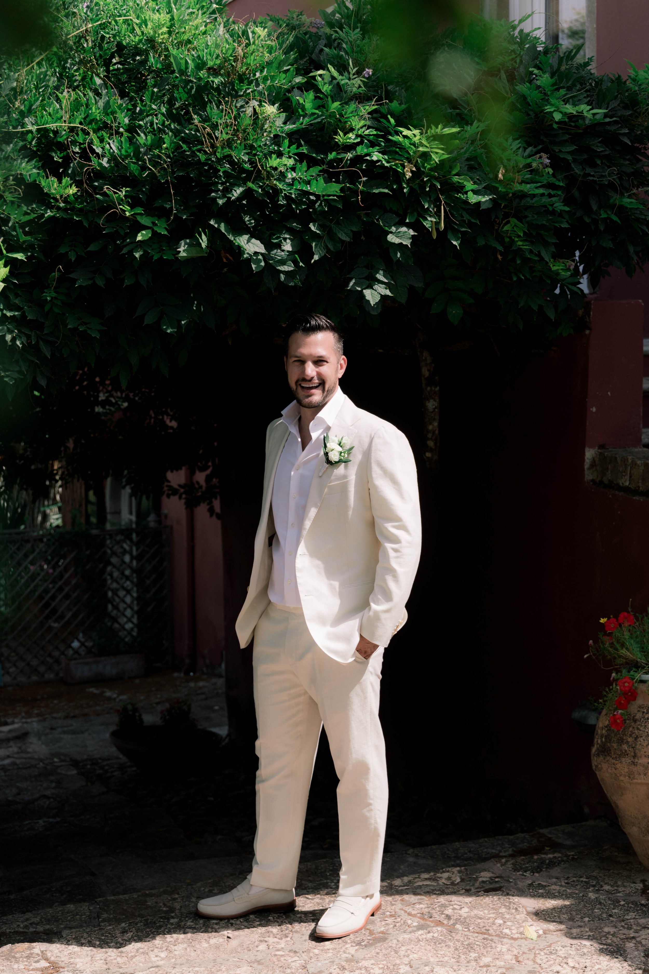 A man in a white suit smiling outdoors with a large leafy green tree behind him.