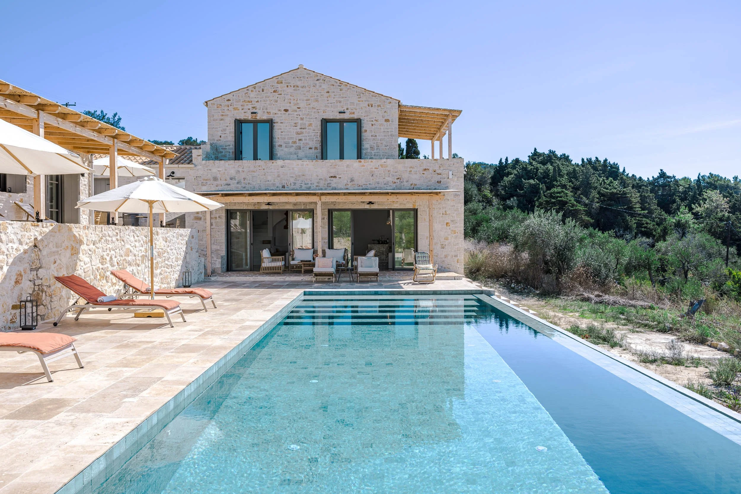 Modern house with a swimming pool in the backyard, sun loungers, umbrellas, and outdoor seating area on a stone patio, surrounded by trees and clear blue sky.