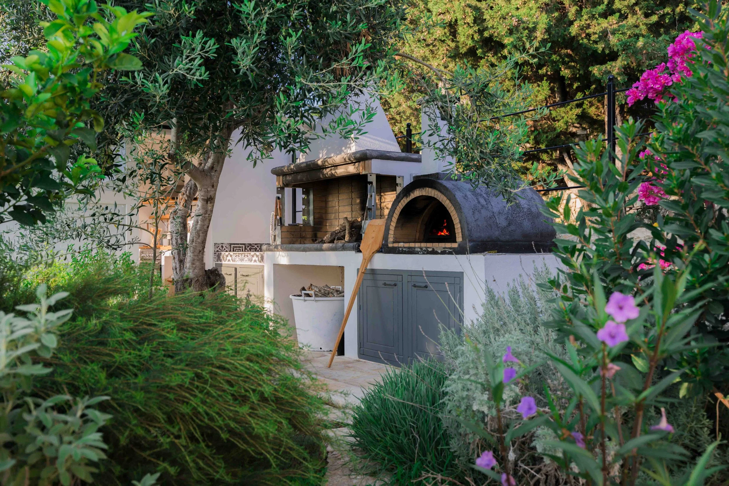 Outdoor brick and concrete pizza oven with fire inside, surrounded by lush green plants and flowers, including purple and pink blossoms.