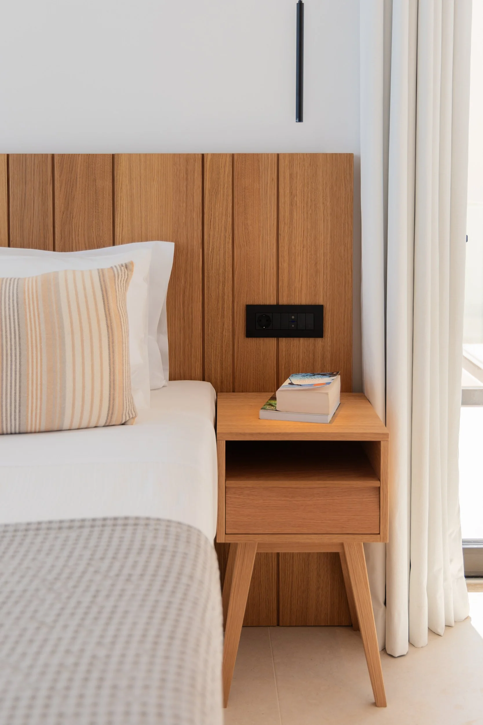 Bedroom with a wooden headboard, a bedside table holding a book and a magazine, and a power outlet on the headboard. White curtains are beside a window.