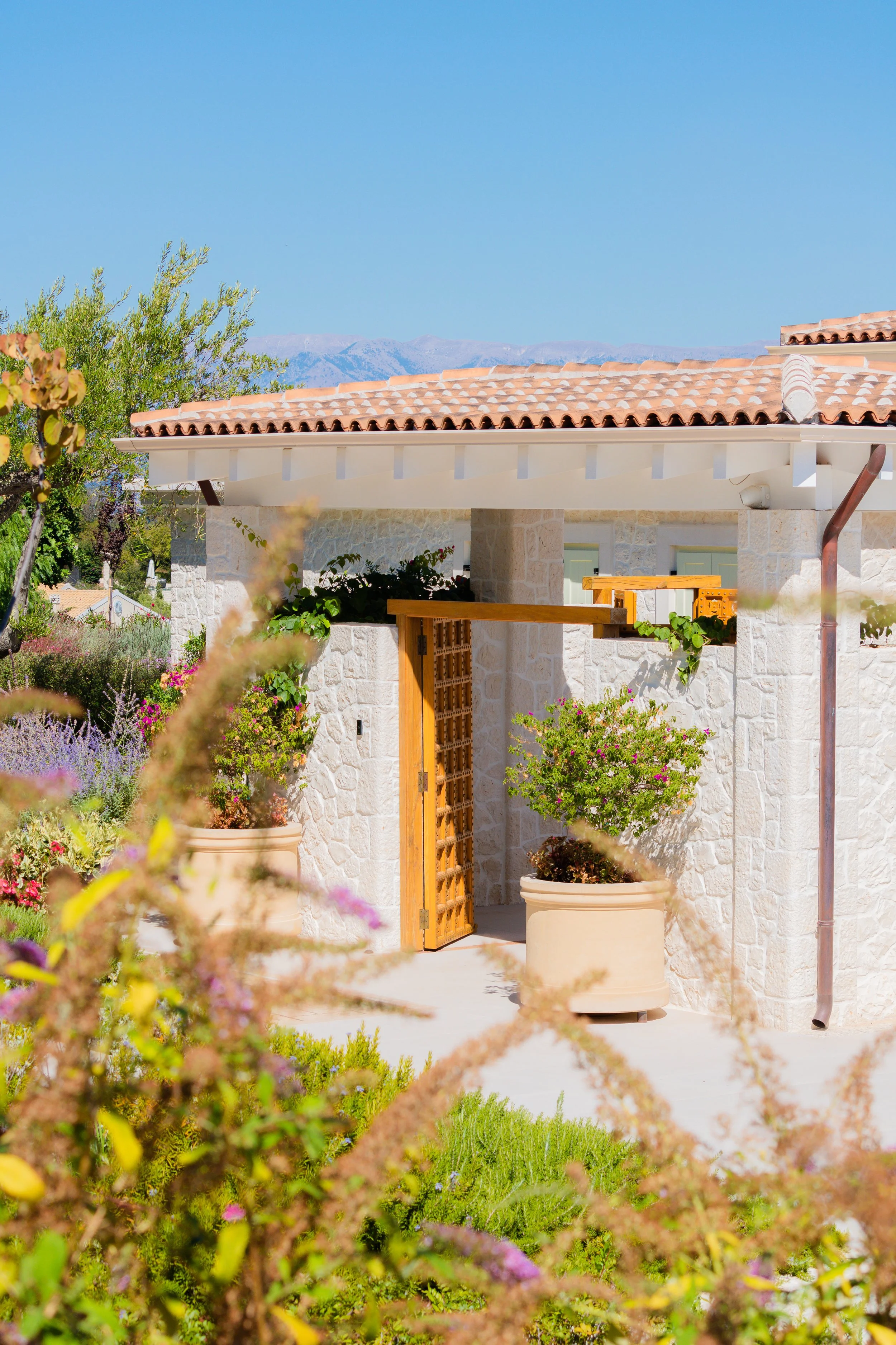 A house with stone walls, a tan tile roof, and a wooden door, surrounded by potted plants and garden flowers with mountains in the background.