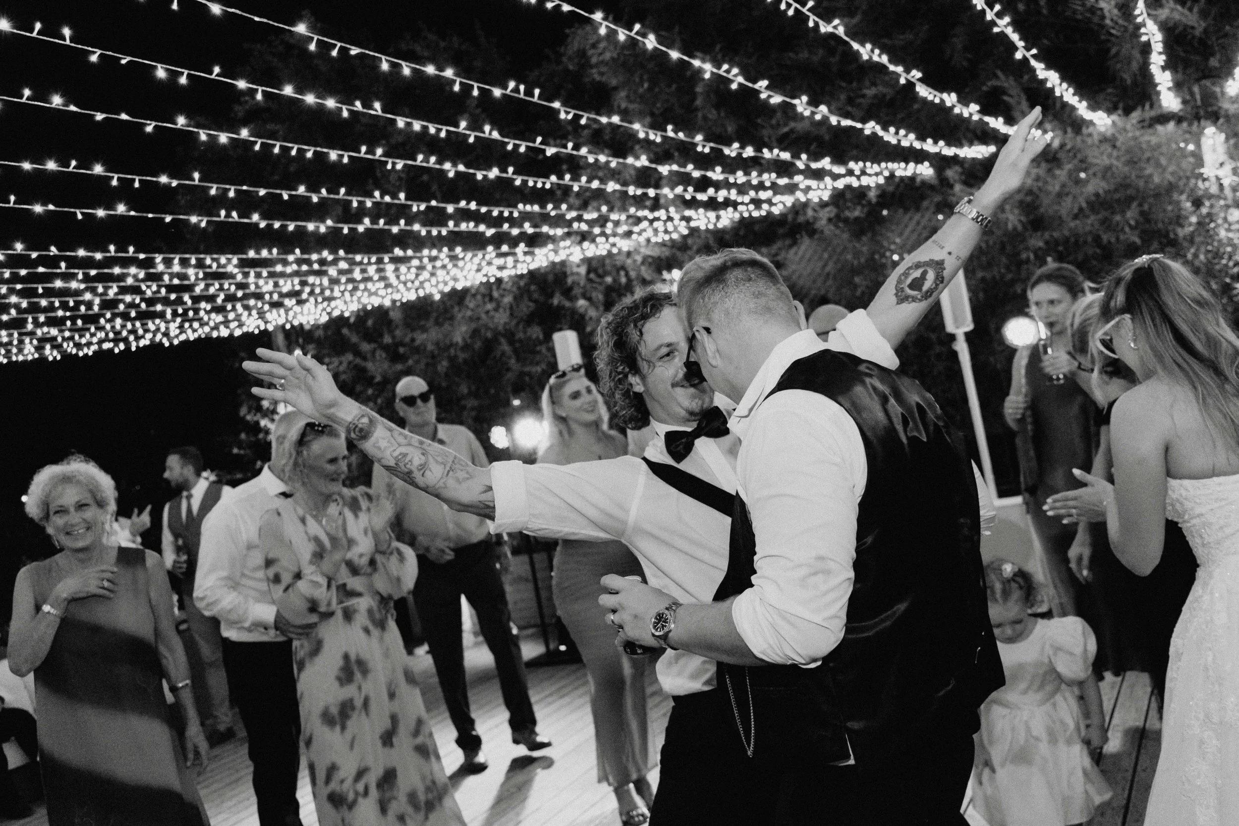 People dancing at a wedding reception under string lights, with some clapping and smiling.