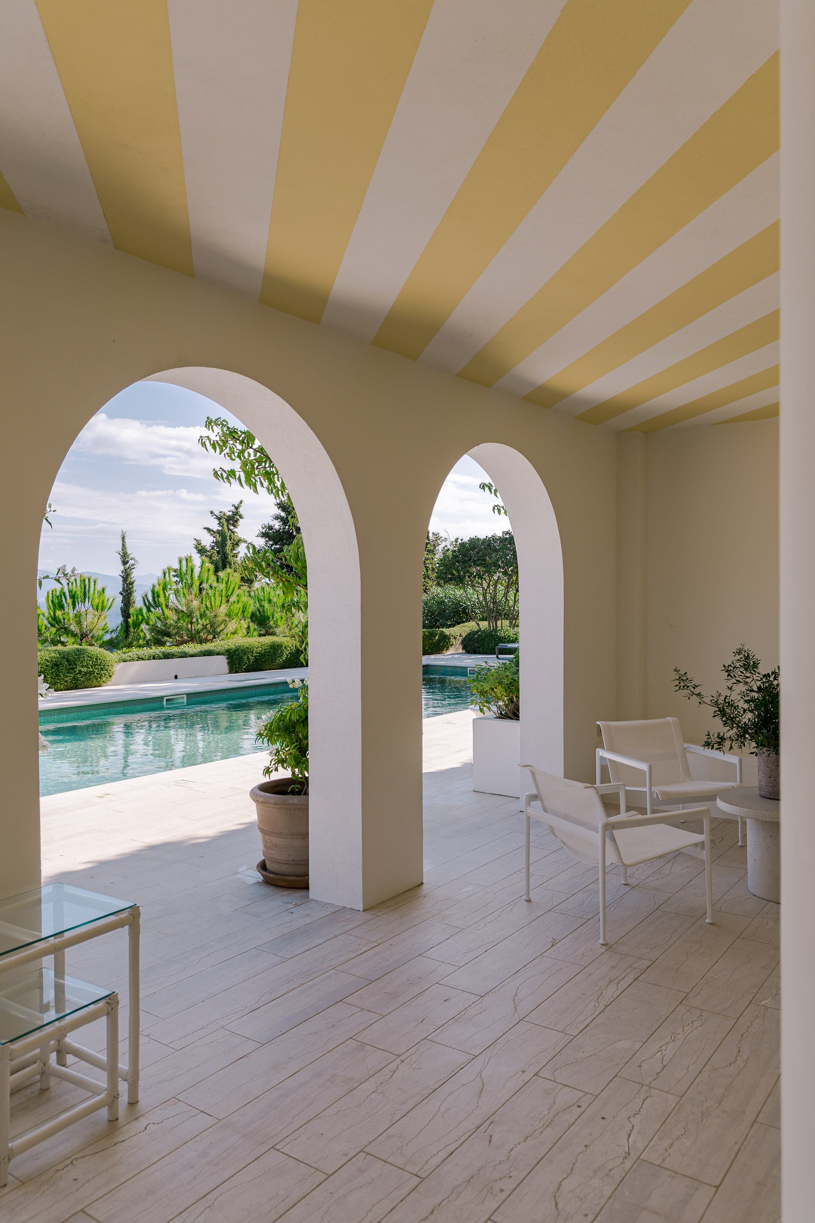 View of a patio or porch area with white chairs and potted plants, overlooking a swimming pool and lush green trees outdoors under a partly cloudy sky.