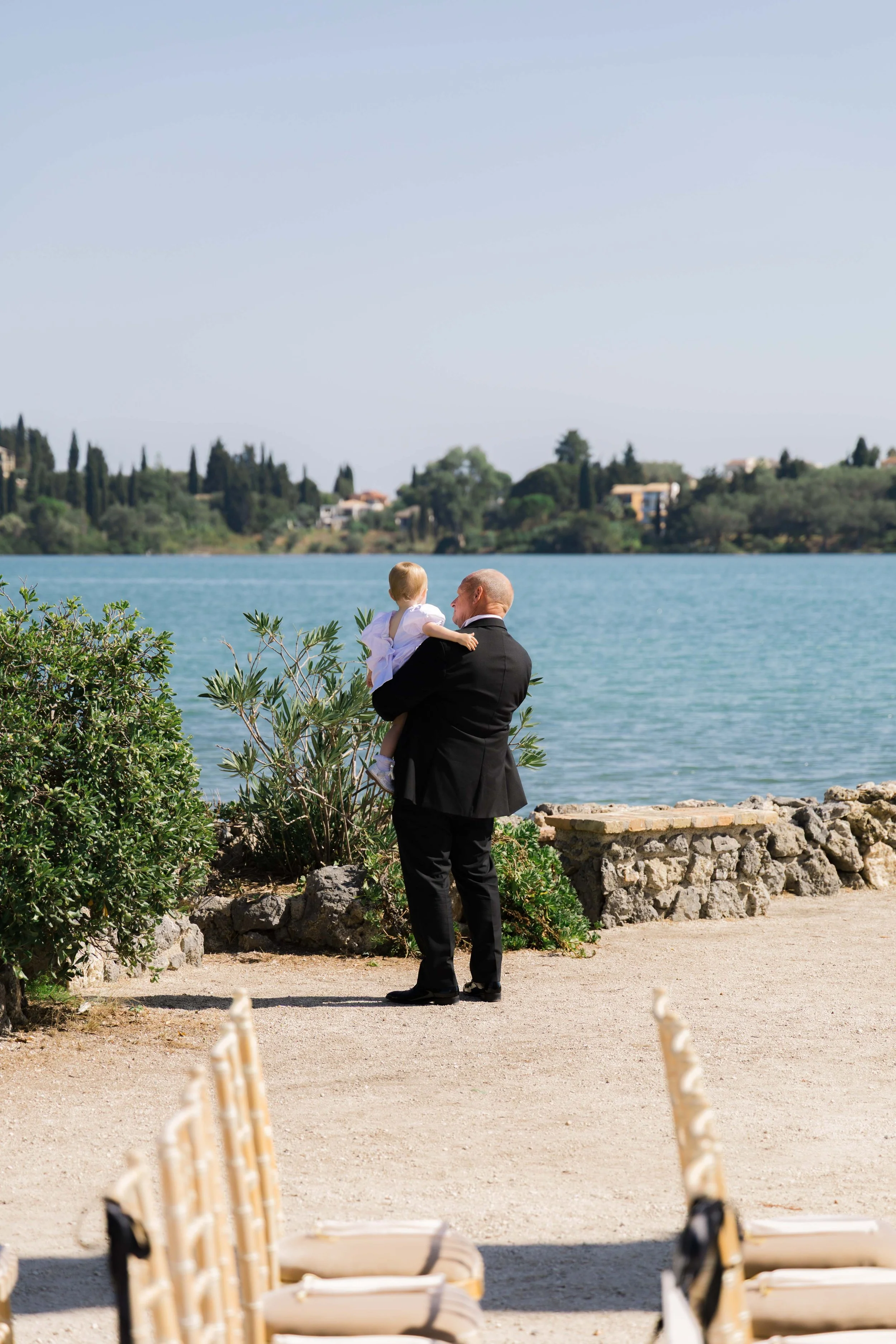 A man in a black suit holding a young girl in white near a body of water on a clear day.