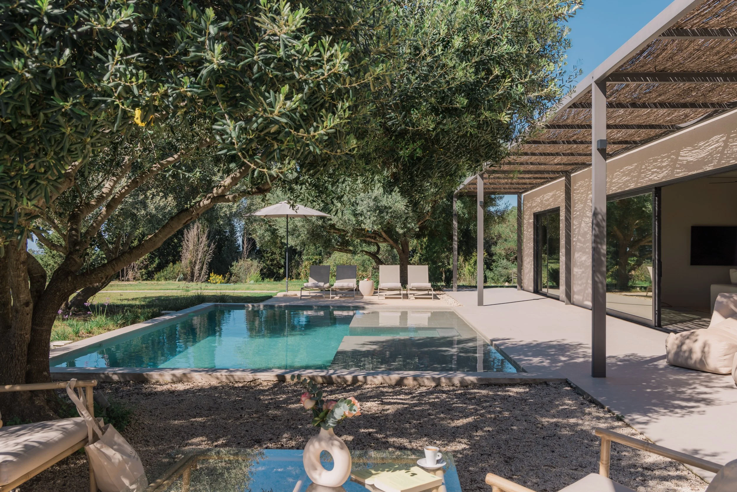 A backyard with a swimming pool, surrounded by trees and lounge chairs, with a patio area attached to a modern house that has sliding glass doors, a with a TV inside visible through the glass.