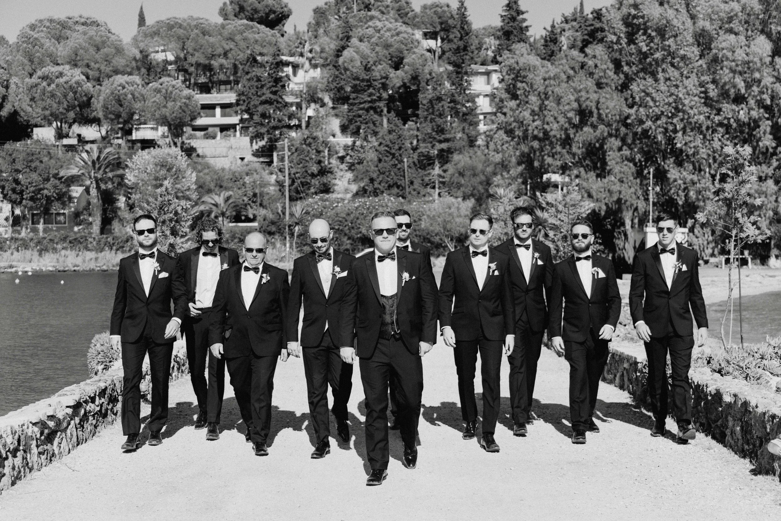 Black and white photo of a group of men walking on a pathway near water, all dressed in formal tuxedos with bow ties and sunglasses, with a background of trees and buildings.