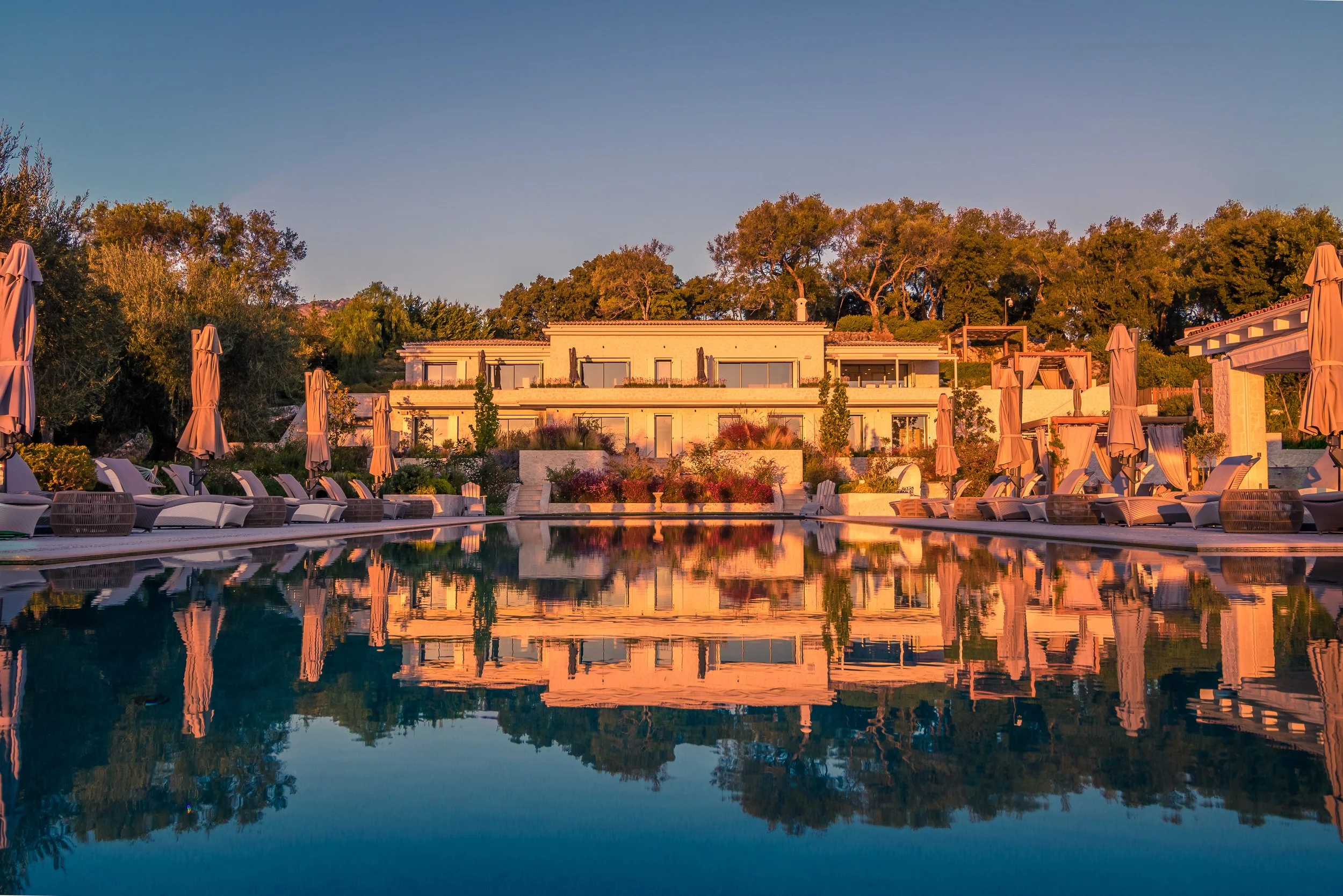 Luxury resort with a large swimming pool, lounge chairs, umbrellas, and a modern building surrounded by trees at sunset.