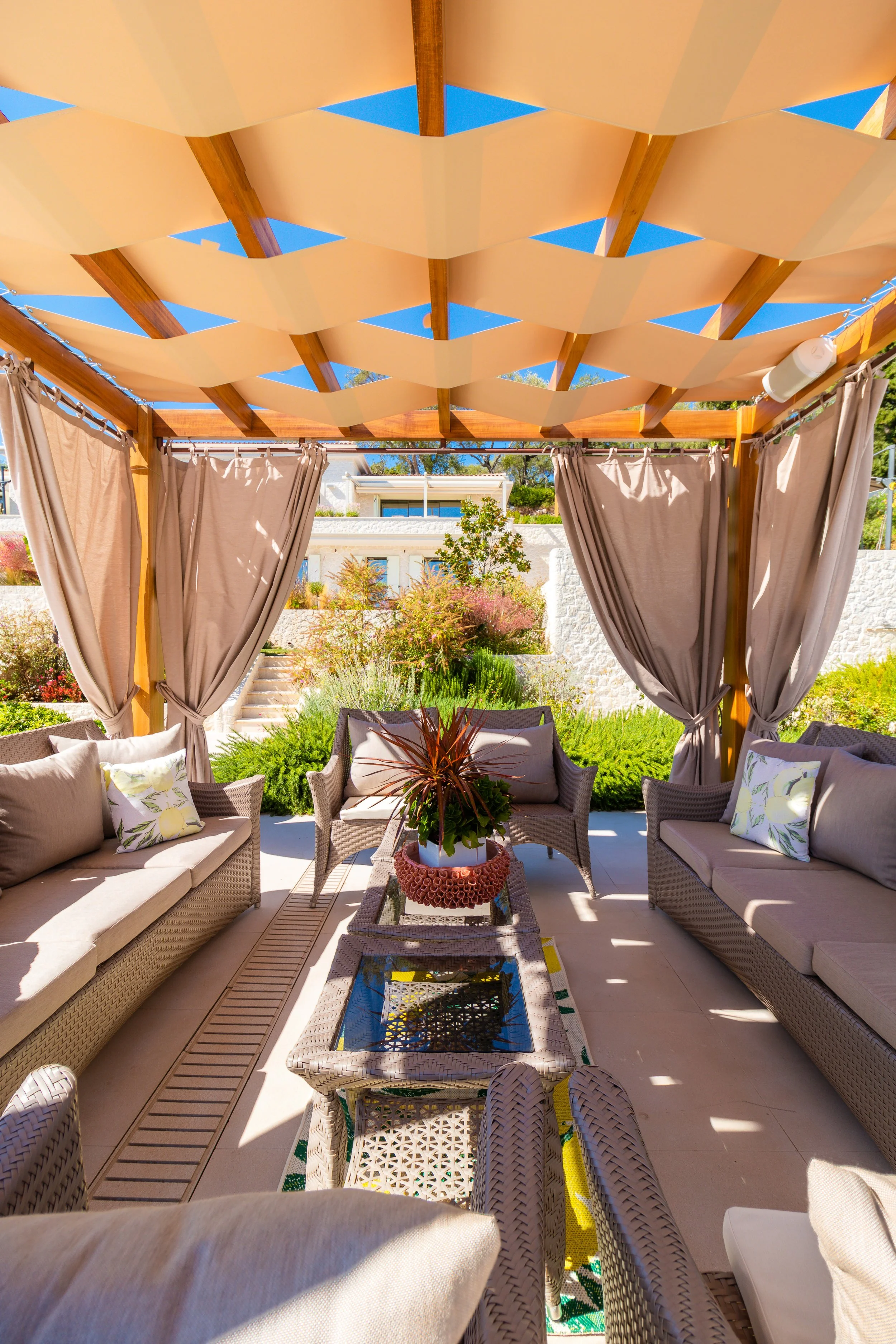 An outdoor patio lounge area with wicker sofas and chairs, beige cushions, a glass-topped coffee table with a potted plant, beige curtains, and a wooden pergola with fabric shading, overlooking a landscaped garden with steps and shrubs.