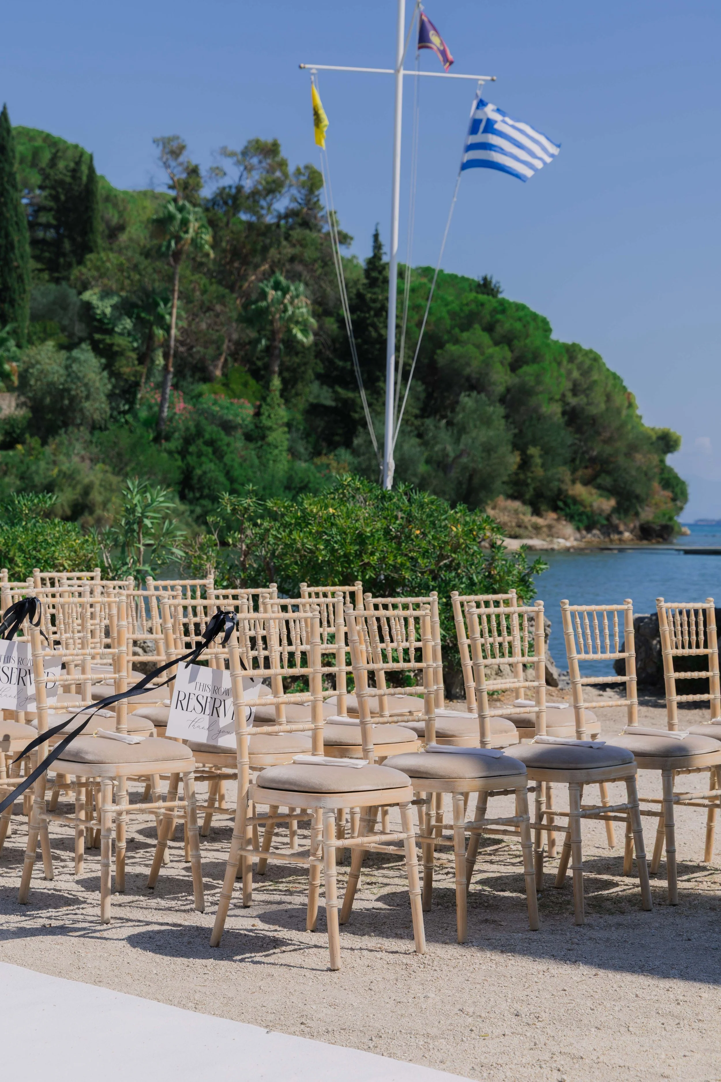 Beachside wedding setup with wooden chairs, reserved signs, flags, and lush greenery near the water.