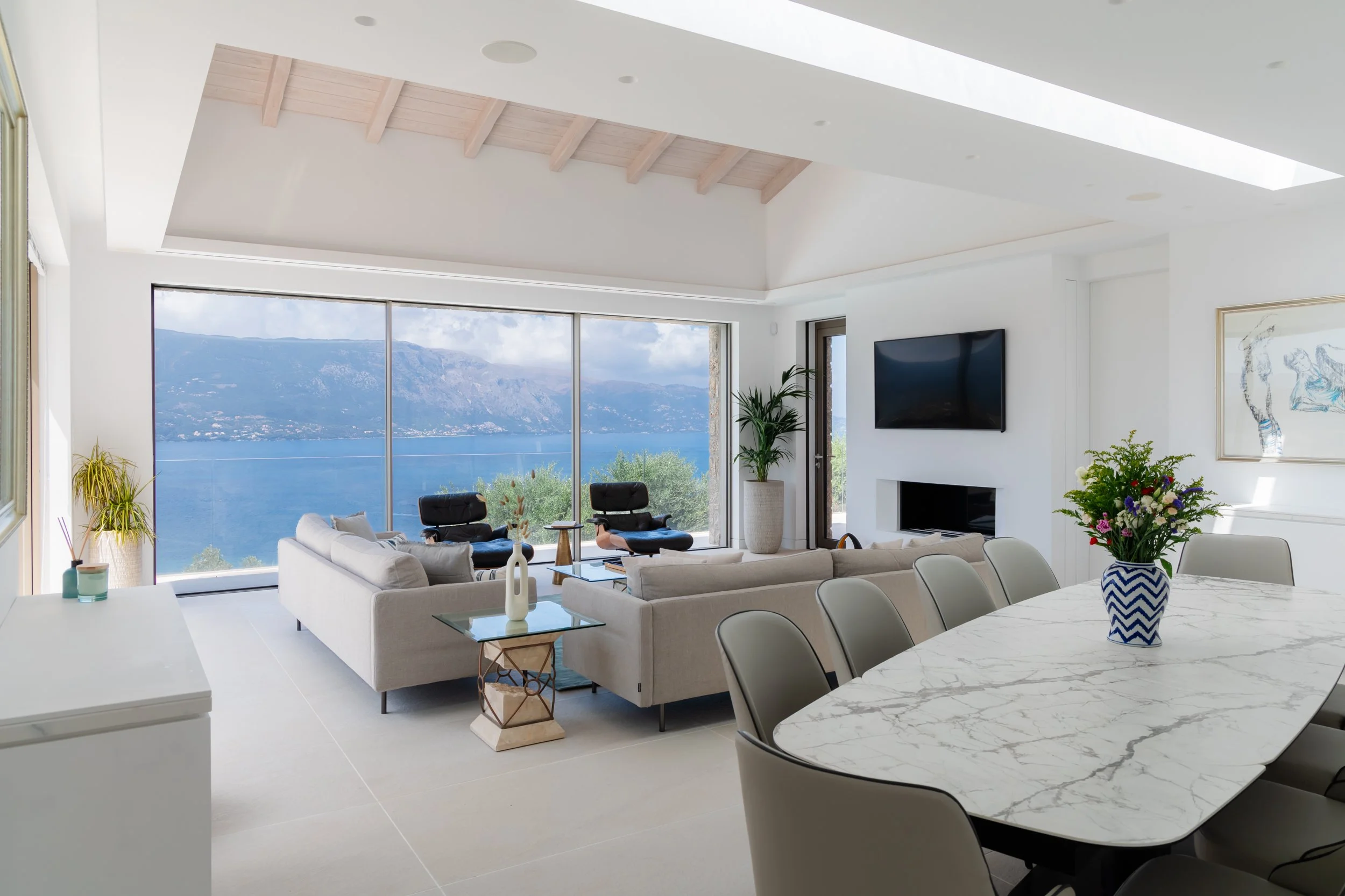 Modern living room with large window overlooking lake and mountains, beige sofas, black chairs, flat-screen TV, contemporary artwork, and a dining table with a floral centerpiece.