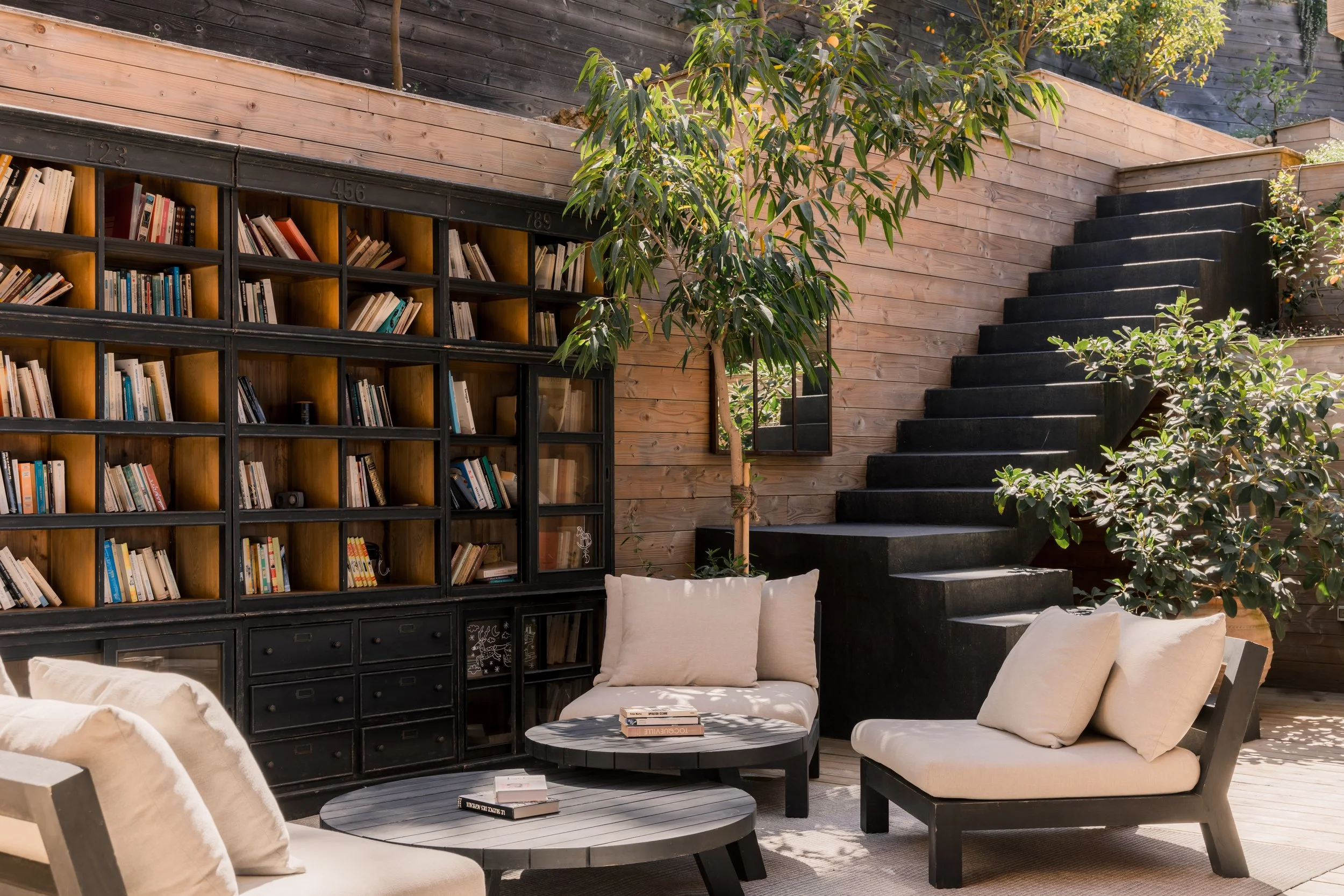 Outdoor patio with black bookshelf filled with books, beige cushioned chairs, a small round black coffee table with books, potted trees, and a black staircase against a wood-paneled wall.