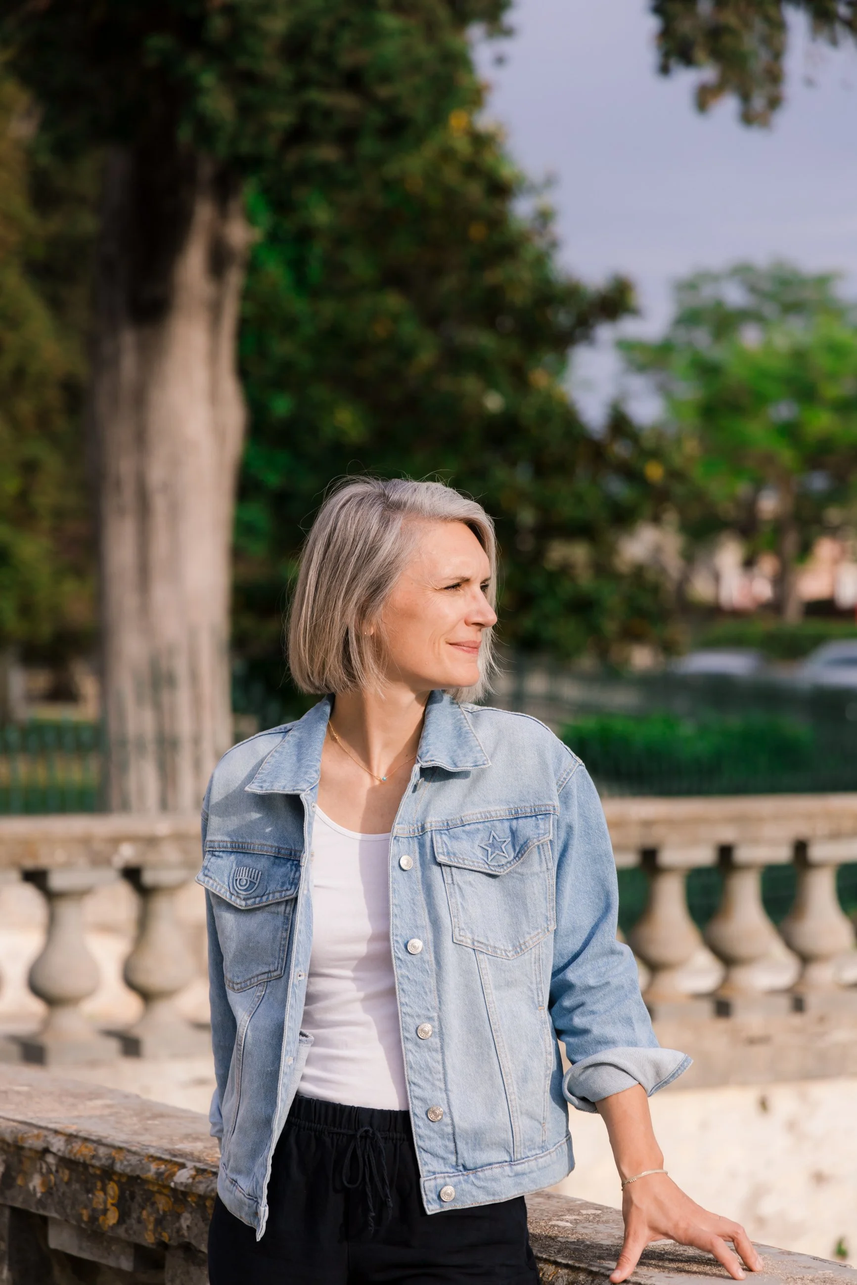 A woman with short gray hair standing outdoors near a stone railing, wearing a denim jacket over a white top, looking to the side with a contemplative expression. Trees and a park-like setting are in the background.