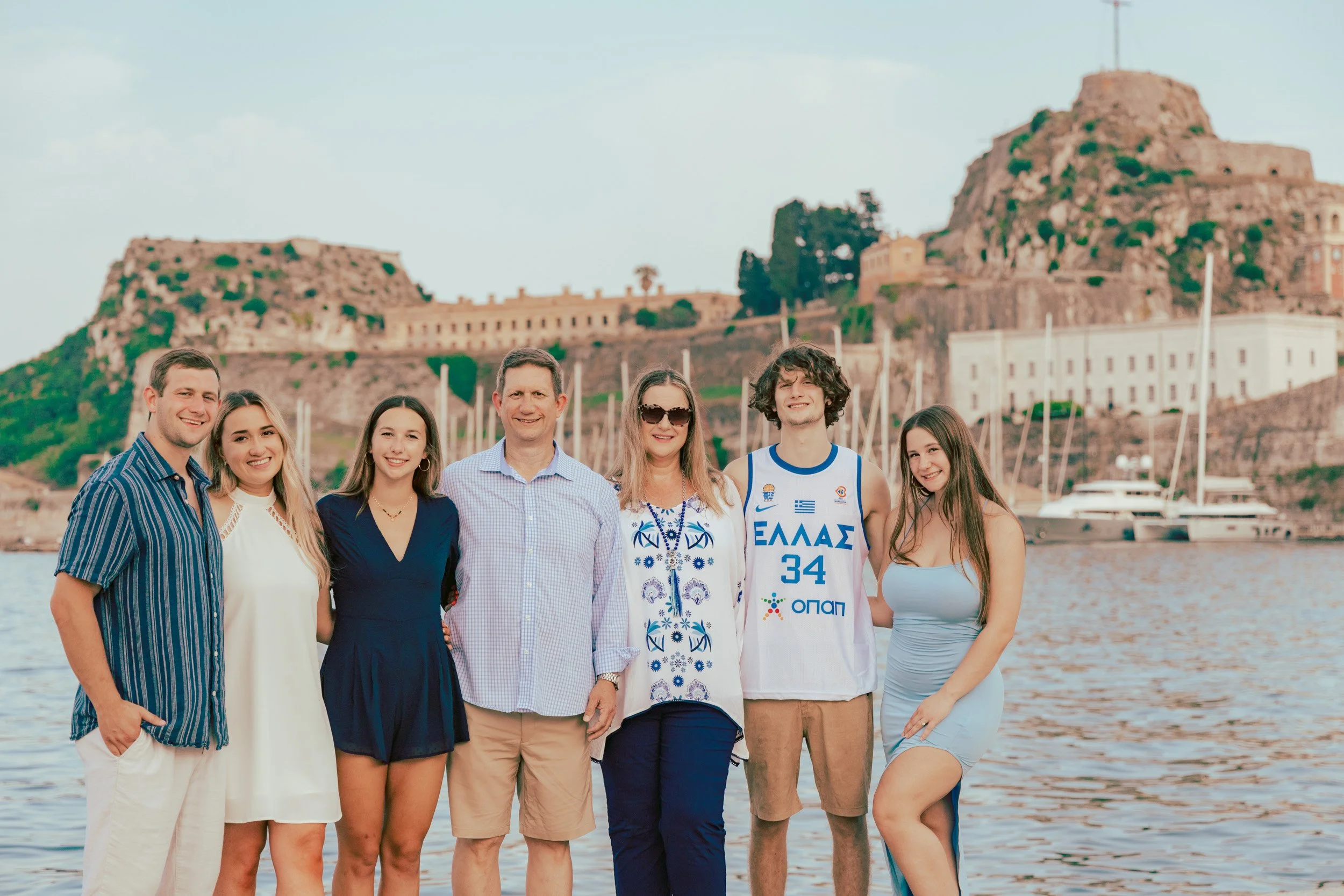 Group of seven people standing together by the water, with a historic fortress on a hill in the background, likely in Greece.