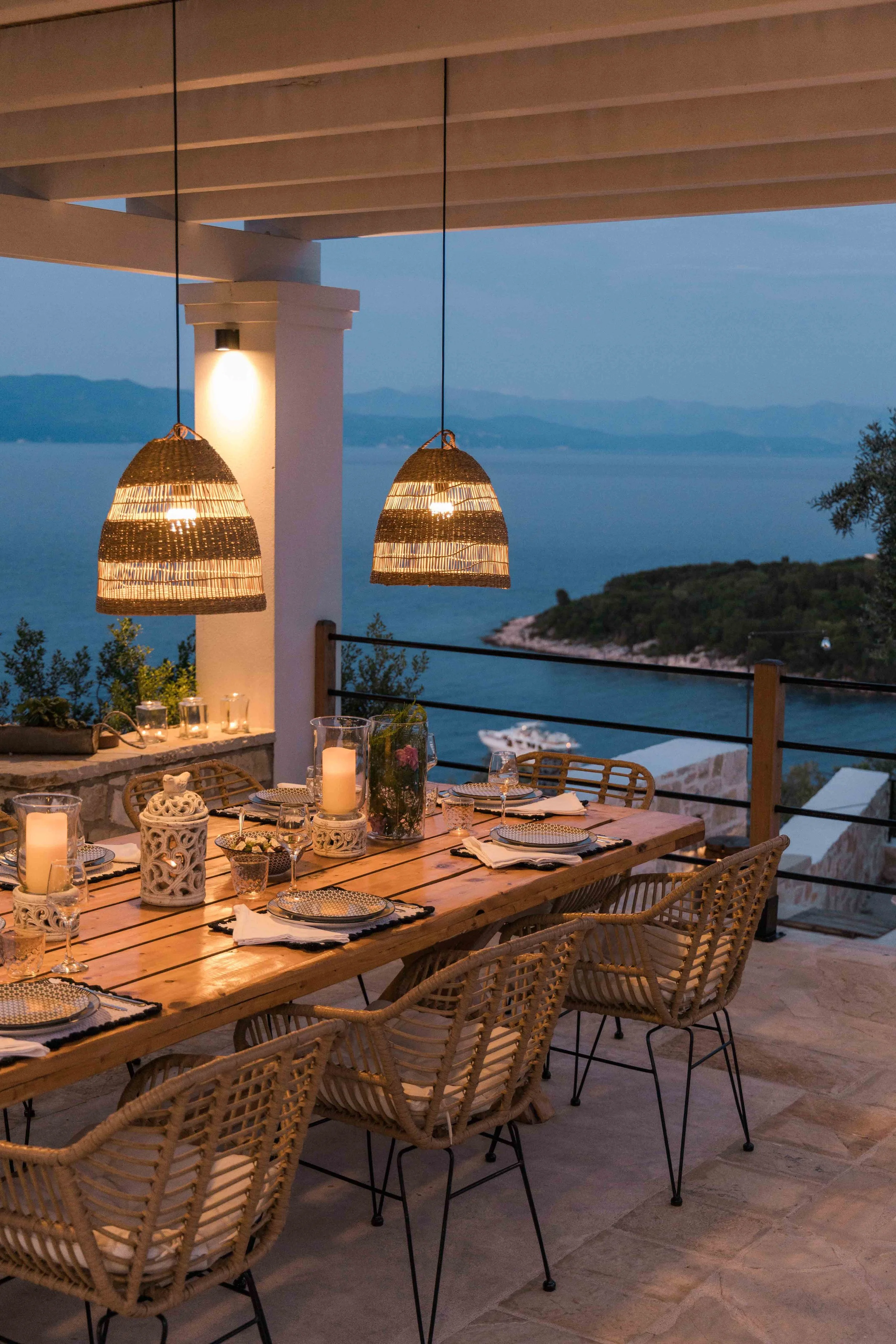 An outdoor dining table set for a meal on a patio overlooking a body of water and distant mountains at dusk. The table features glassware, plates, napkins, candles, and decorative lanterns with hanging wicker light fixtures above.