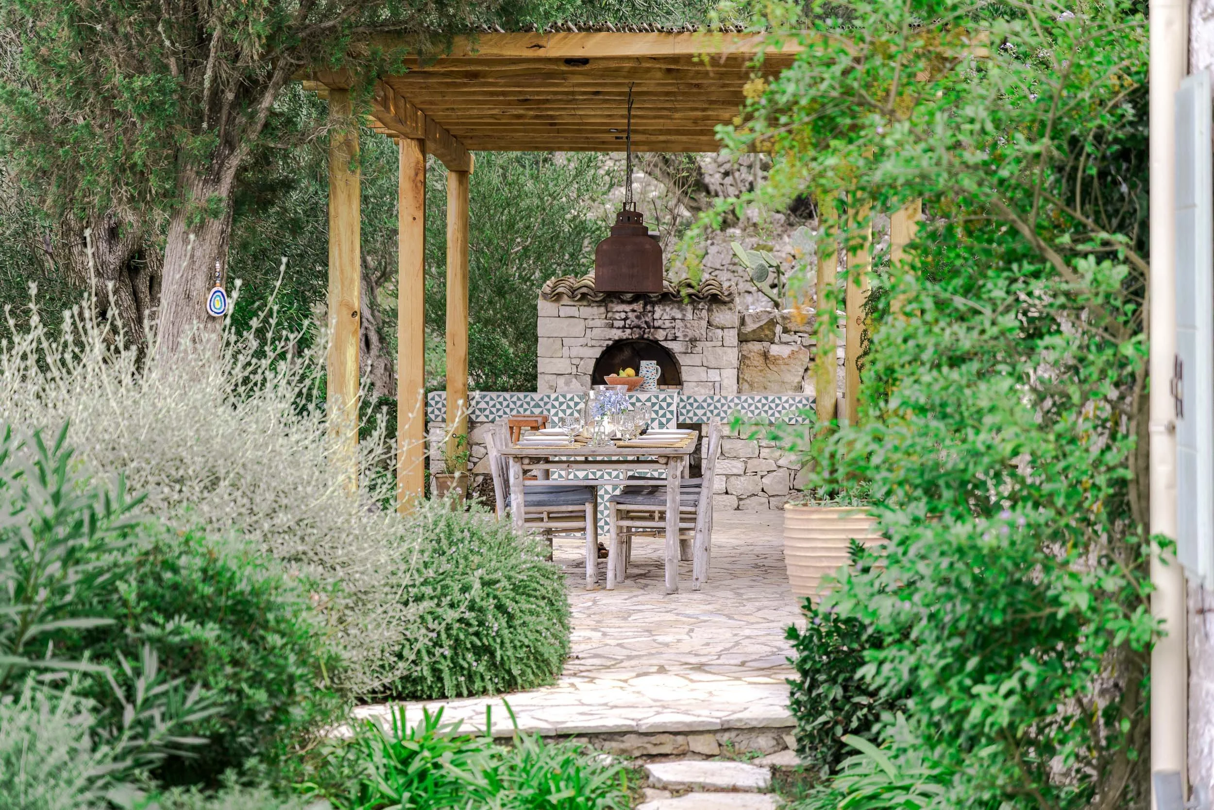 Outdoor dining area with a rustic wooden table and chairs, a stone BBQ or fireplace, surrounded by greenery and a pergola with a hanging light.