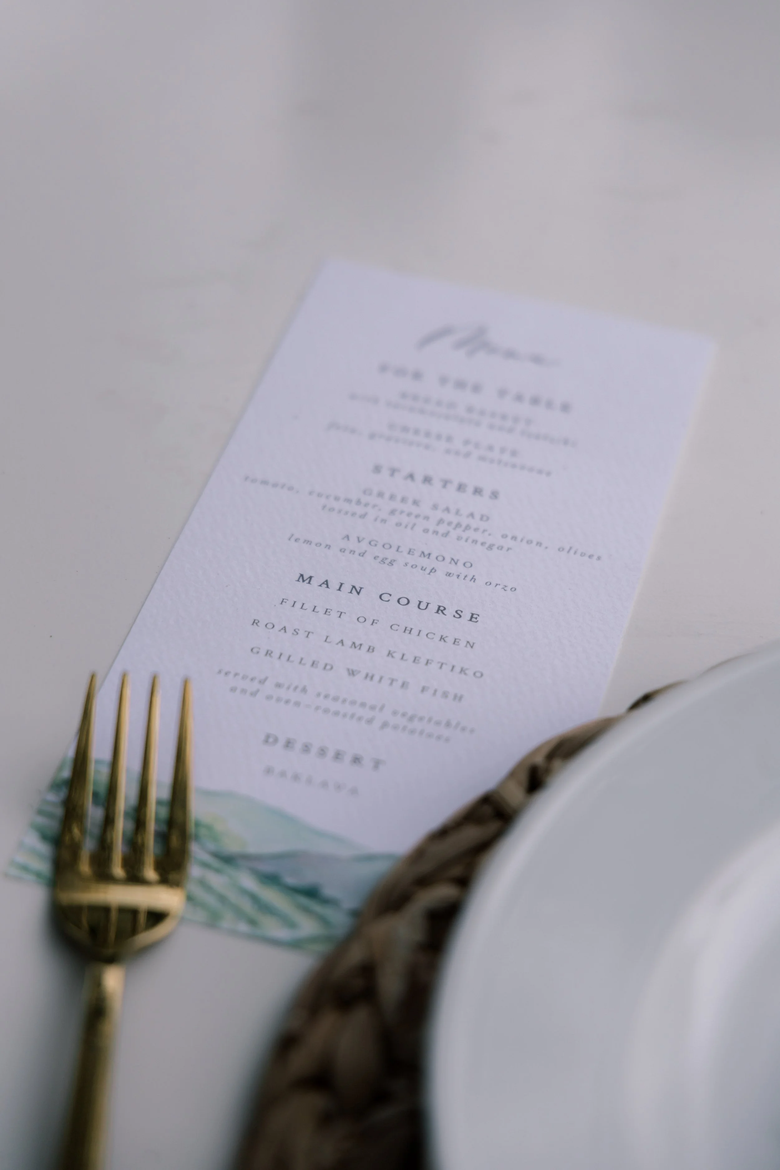 A printed menu placed on a table with a gold fork and a white plate partially visible. The menu has a watercolor illustration at the bottom.