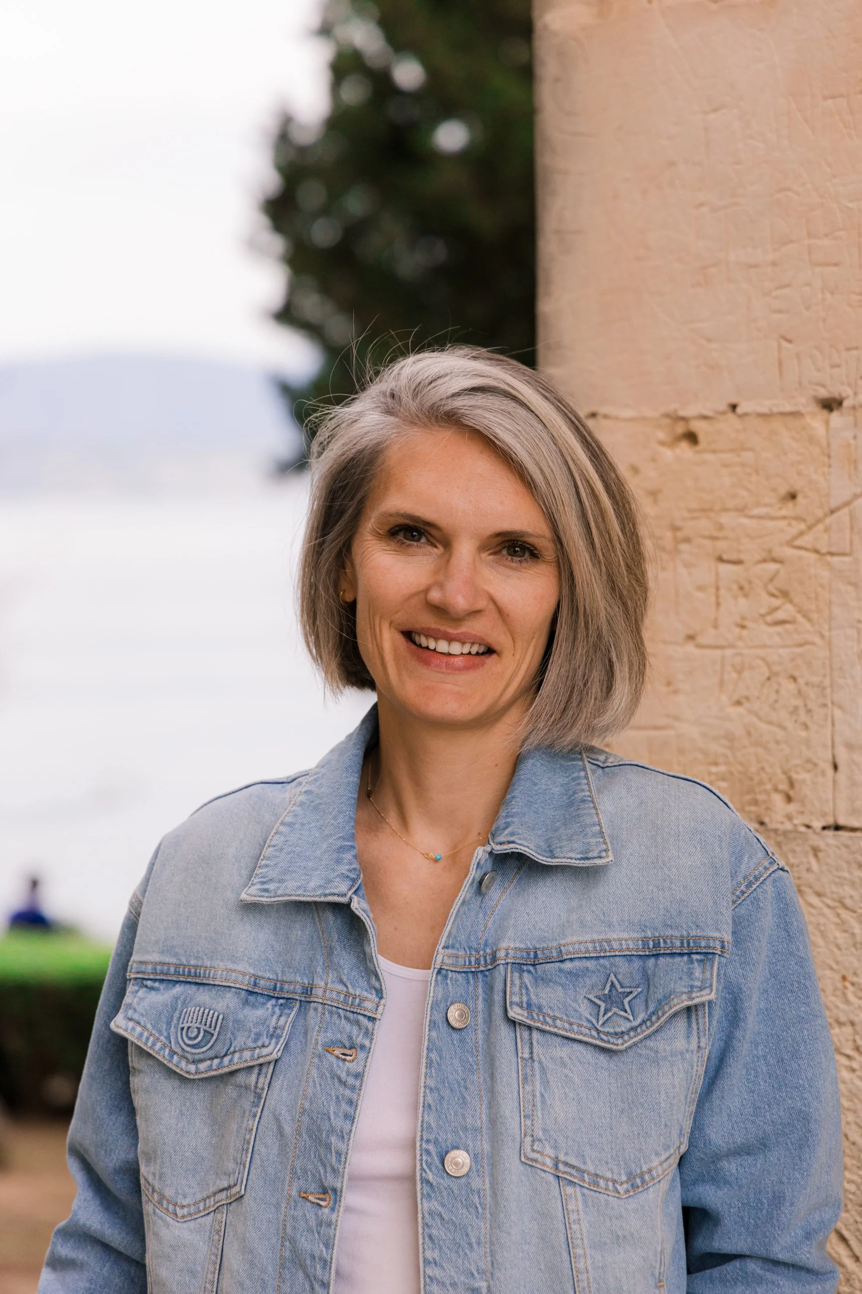 A woman with short gray hair smiling outdoors near stone columns and a blurred background of trees and water.