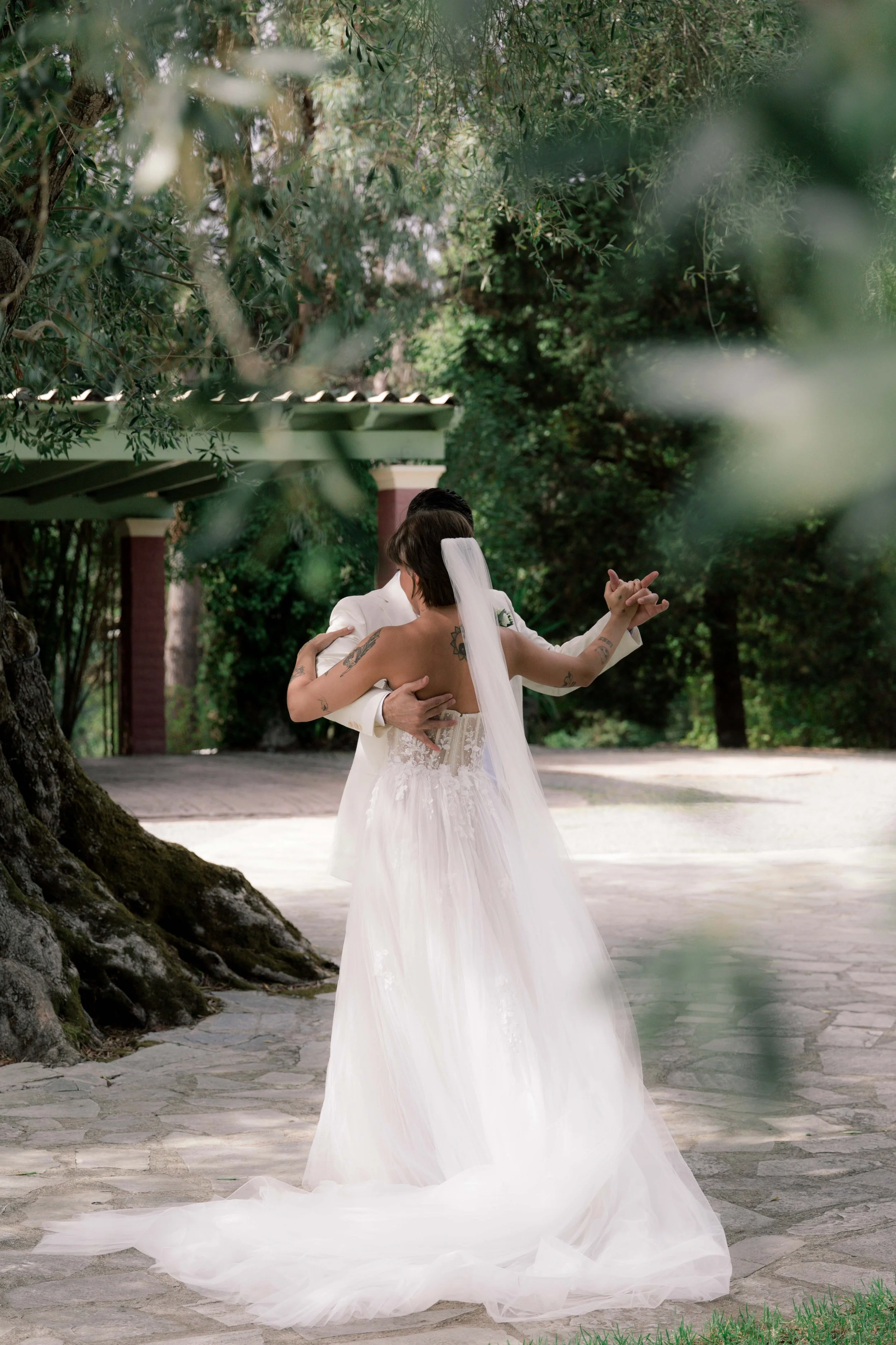 A bride and groom sharing a dance outdoors under trees, with the bride wearing a white wedding gown and veil.