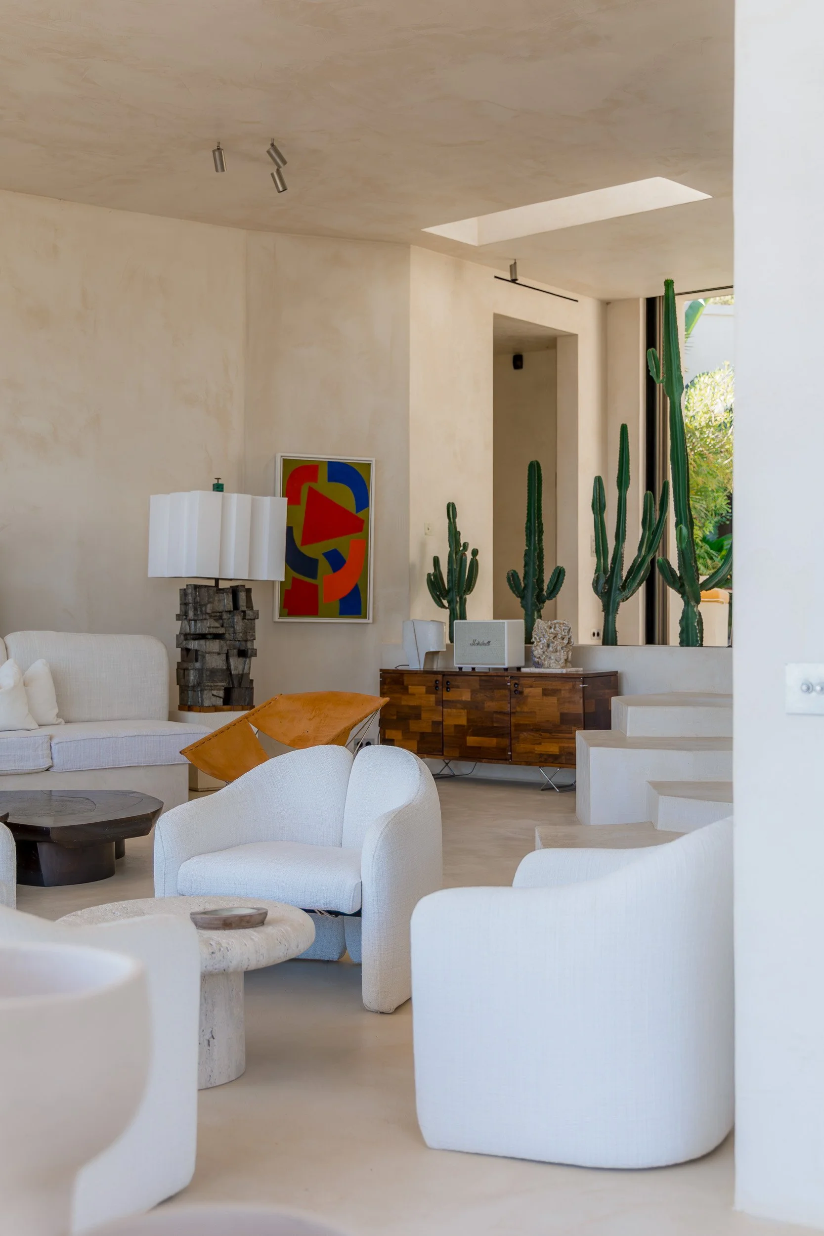 Interior of a modern living room with white chairs, a wooden sideboard, tall cacti, a colorful abstract painting, and a table lamp, with natural light coming through large windows.