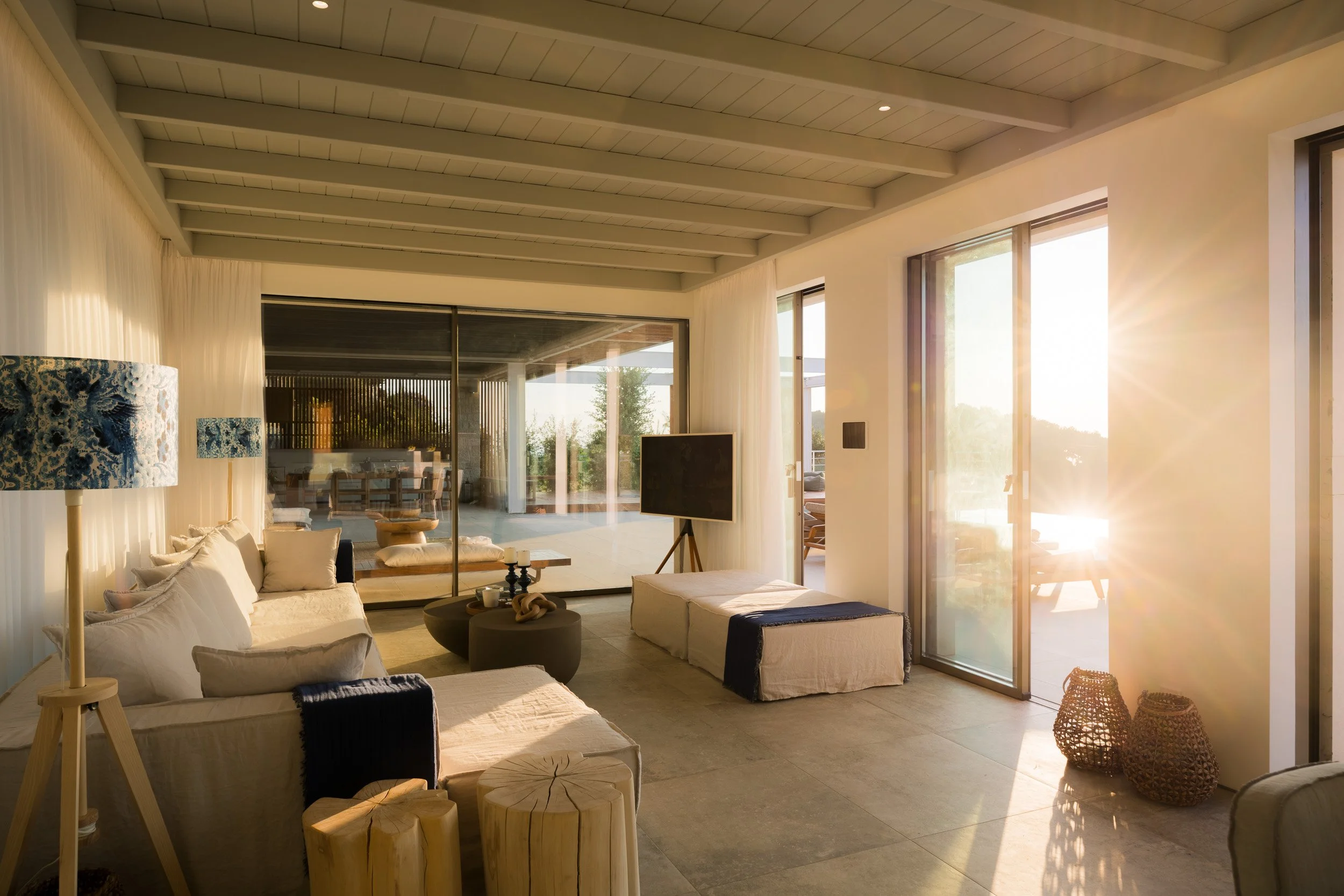Living room with white couches, wooden tables, a TV, sliding glass doors, and sunlight streaming in.