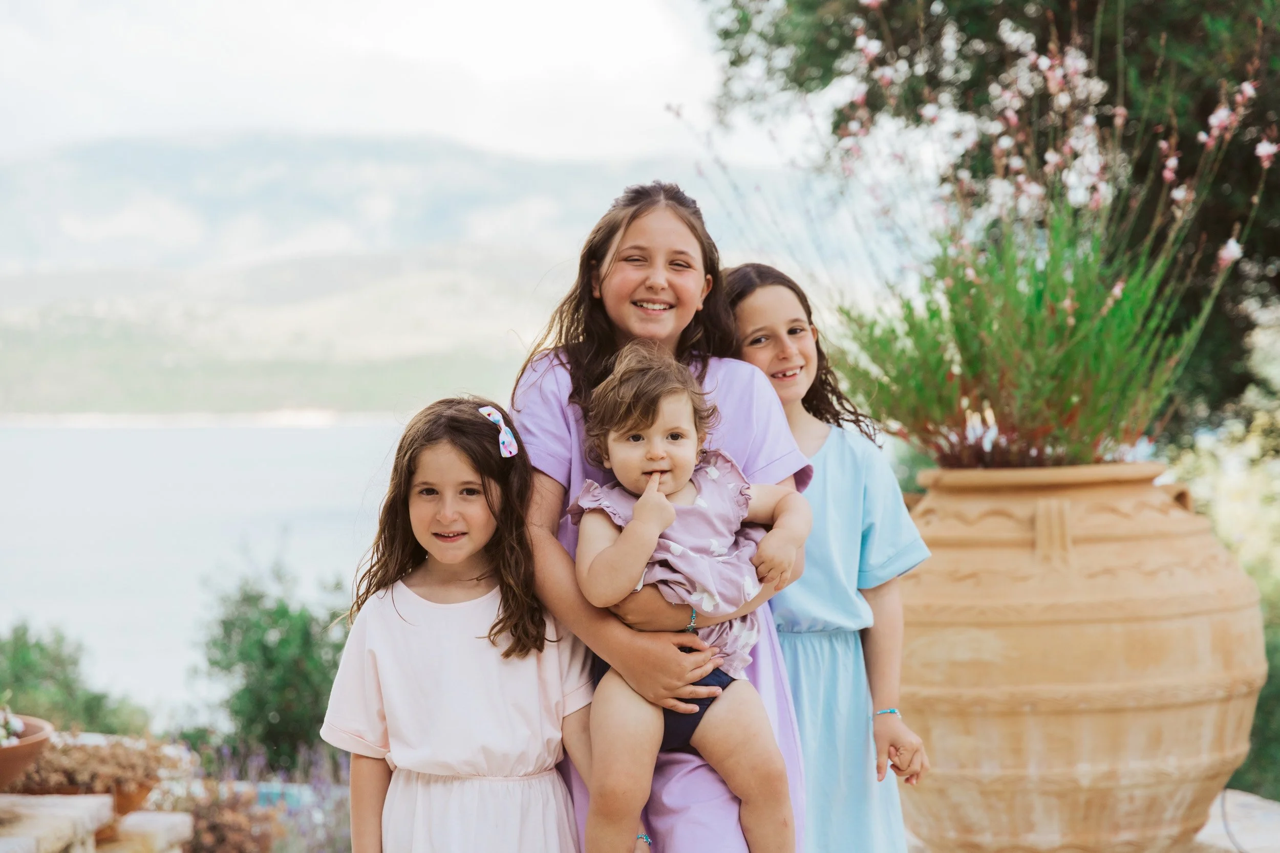 A woman and four young girls pose outdoors near a large decorative pot with plants, with a body of water and mountains in the background.