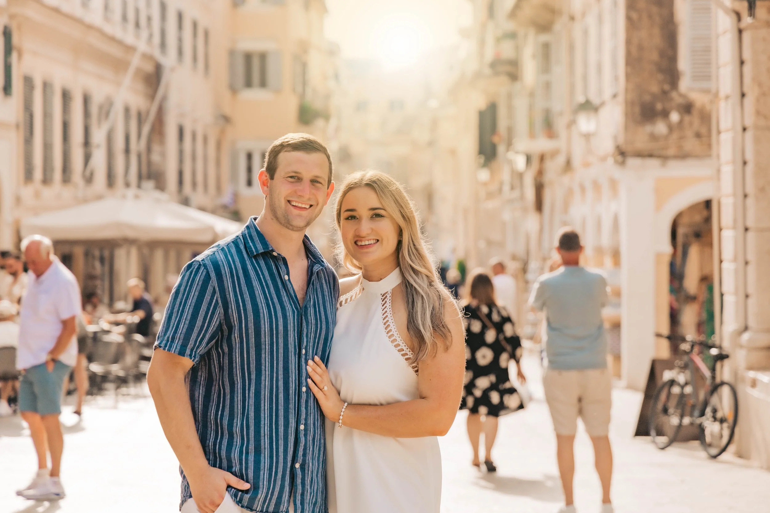 A smiling couple standing close together on a city street on a sunny day with people walking in the background.