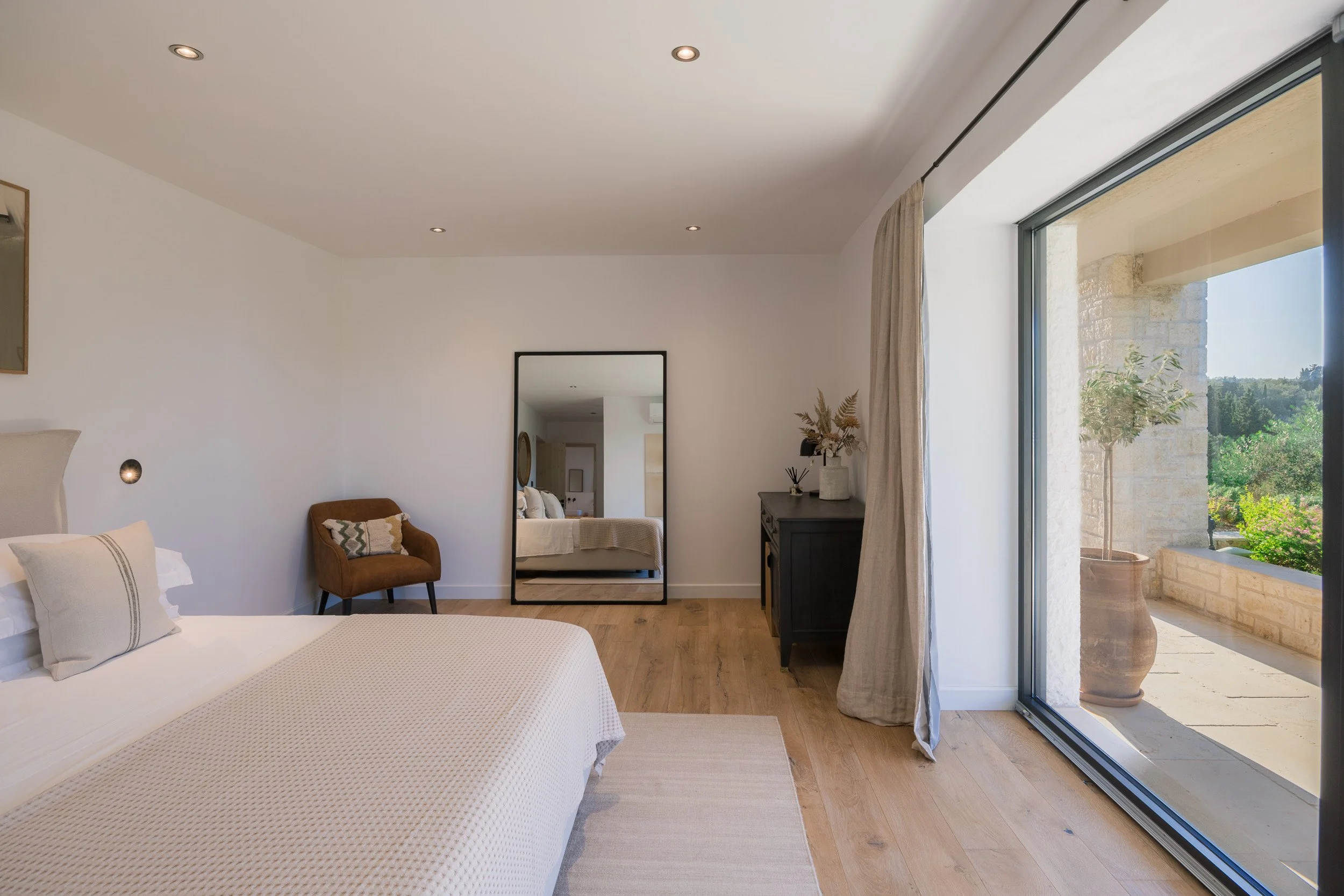 Bright bedroom with a large bed, a mirror, a small brown chair, and a glass door leading to a balcony with a stone wall and potted plant, natural light flooding the room.