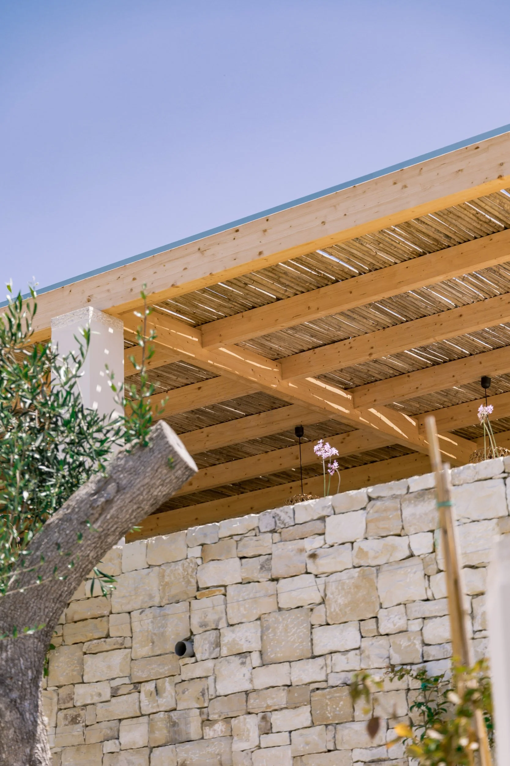 View of an outdoor construction area with a stone wall, wooden beams, and a bamboo roof, under a clear blue sky.
