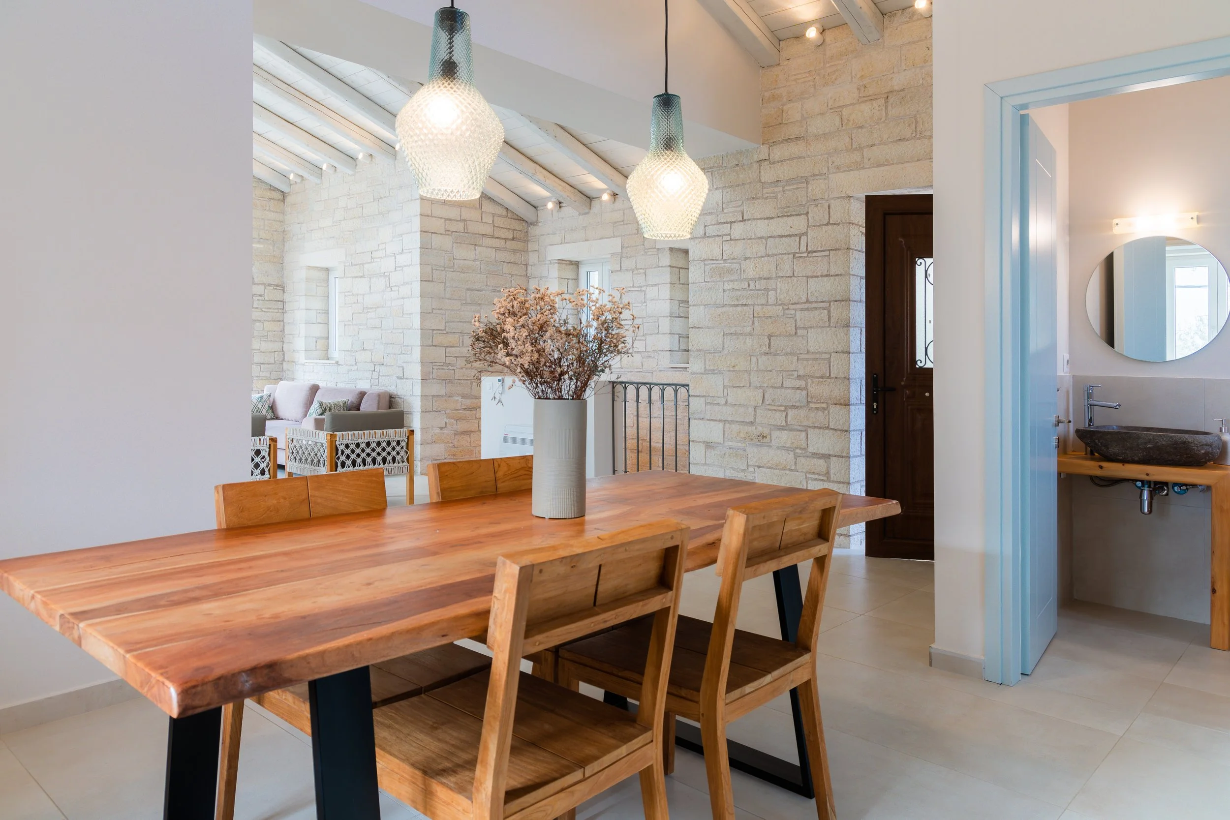 Dining room with wooden table and chairs, decorative vase with dried flowers, pendant lights, exposed brick wall, adjacent bathroom with circular mirror and vessel sink.