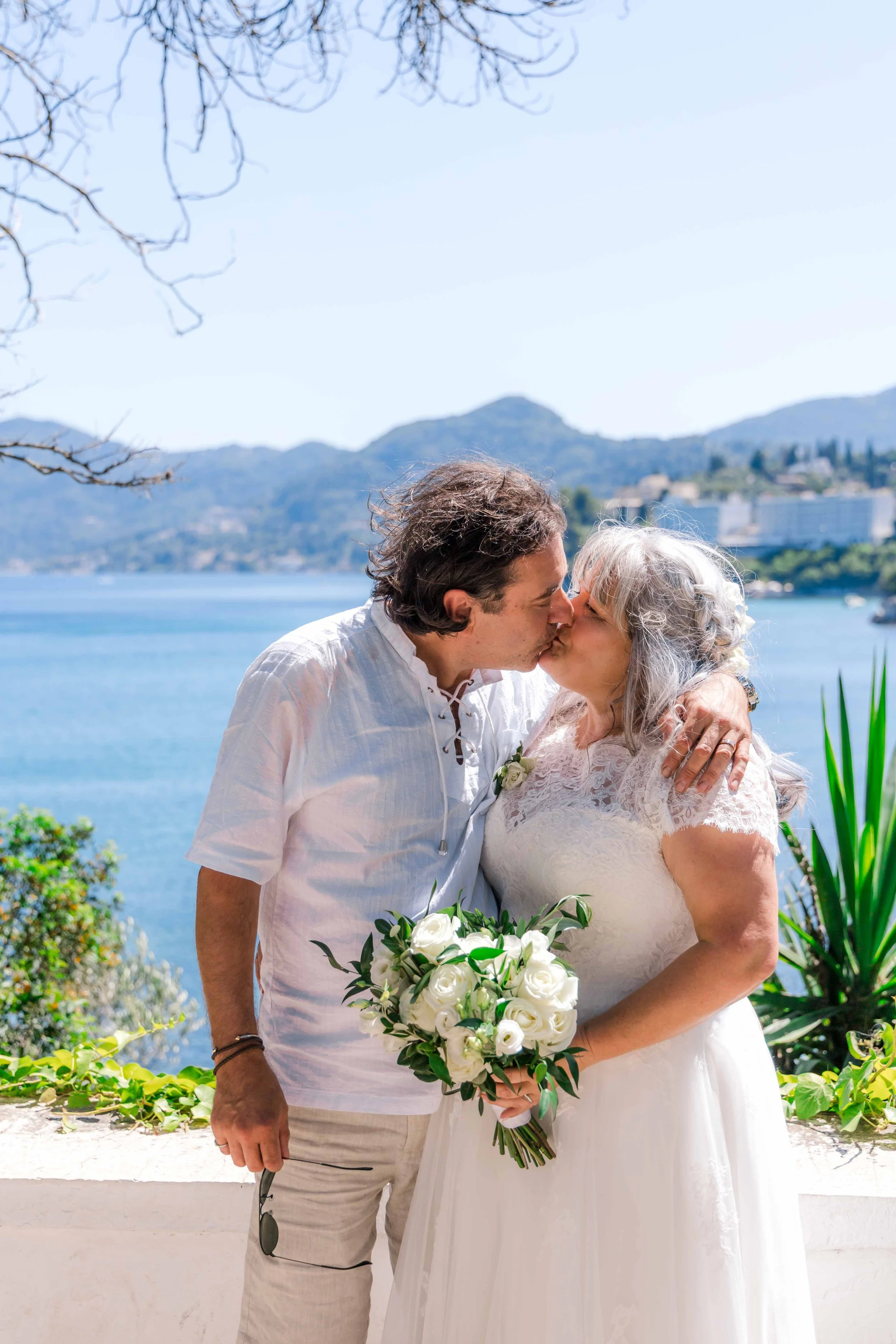 A couple is kissing by a lake, with mountains and buildings in the background, during a wedding or special event.