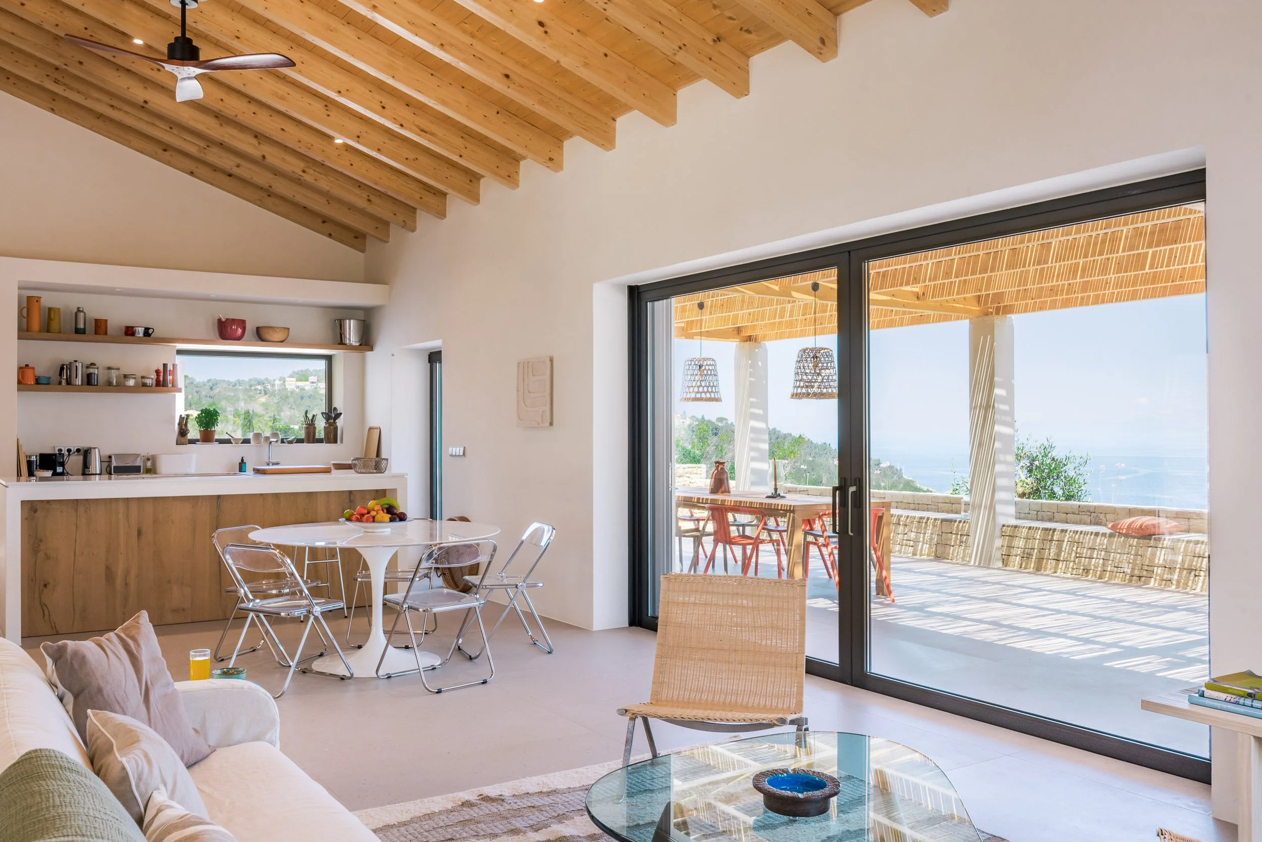 Open-concept living space with a wooden ceiling, large sliding glass doors leading to an outdoor patio, a dining table with chairs, a kitchen with open shelving, and glimpses of a scenic view of greenery and ocean outside.