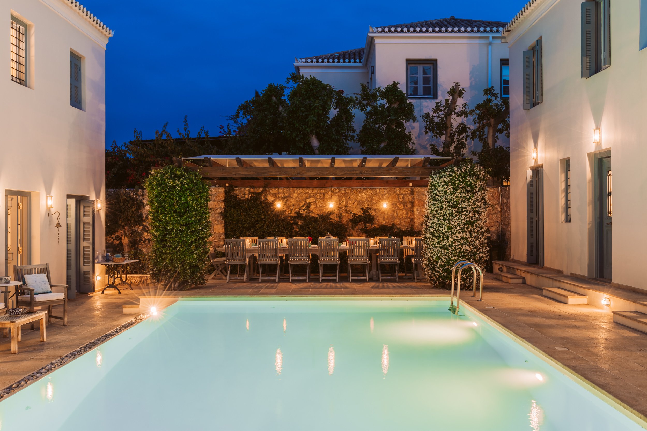 A backyard with a swimming pool, outdoor seating, string lights, and a dining table under a pergola at dusk.