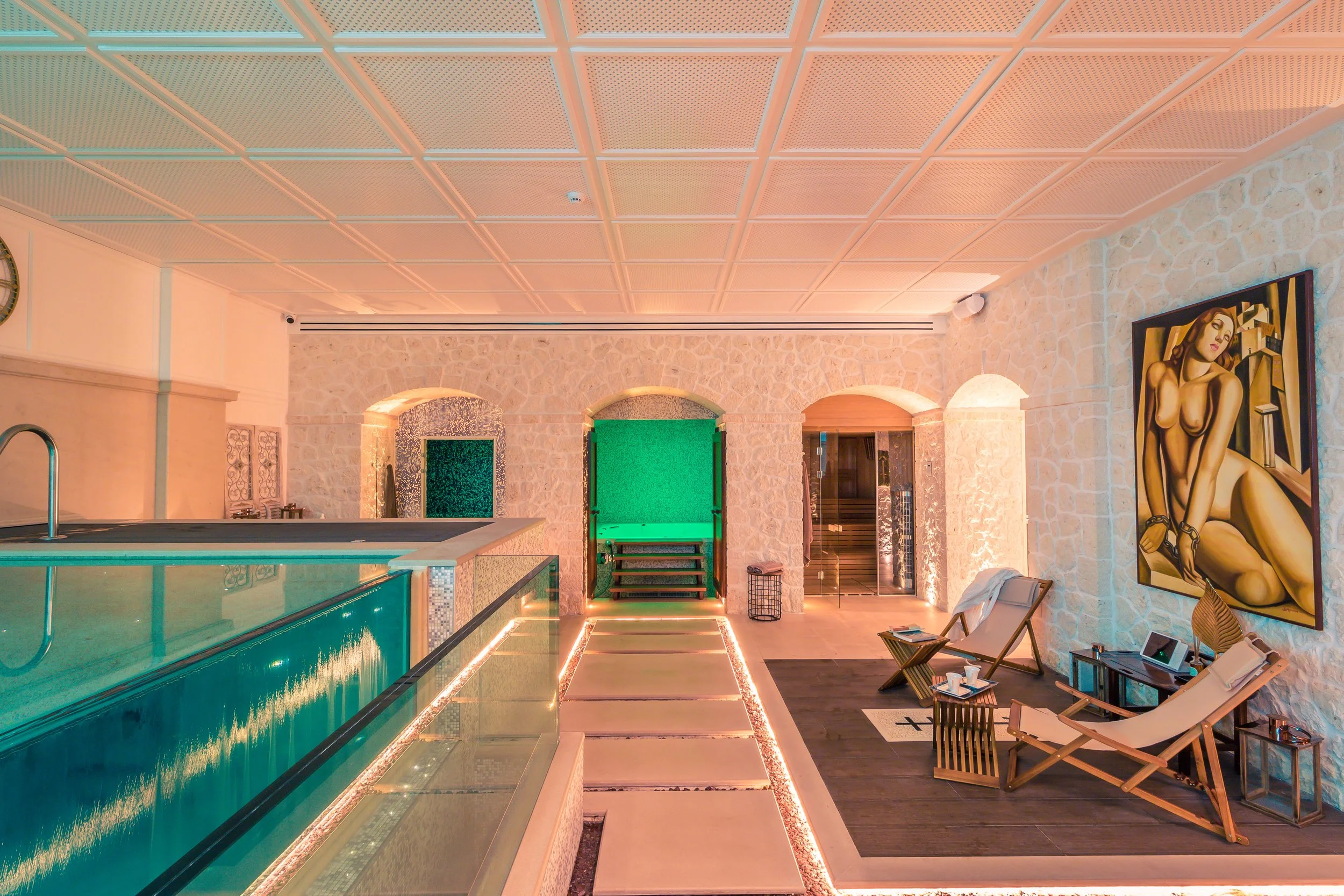 Interior of a luxurious spa or wellness center with a small pool, lounge chairs, a colorful abstract painting, and stone walls.