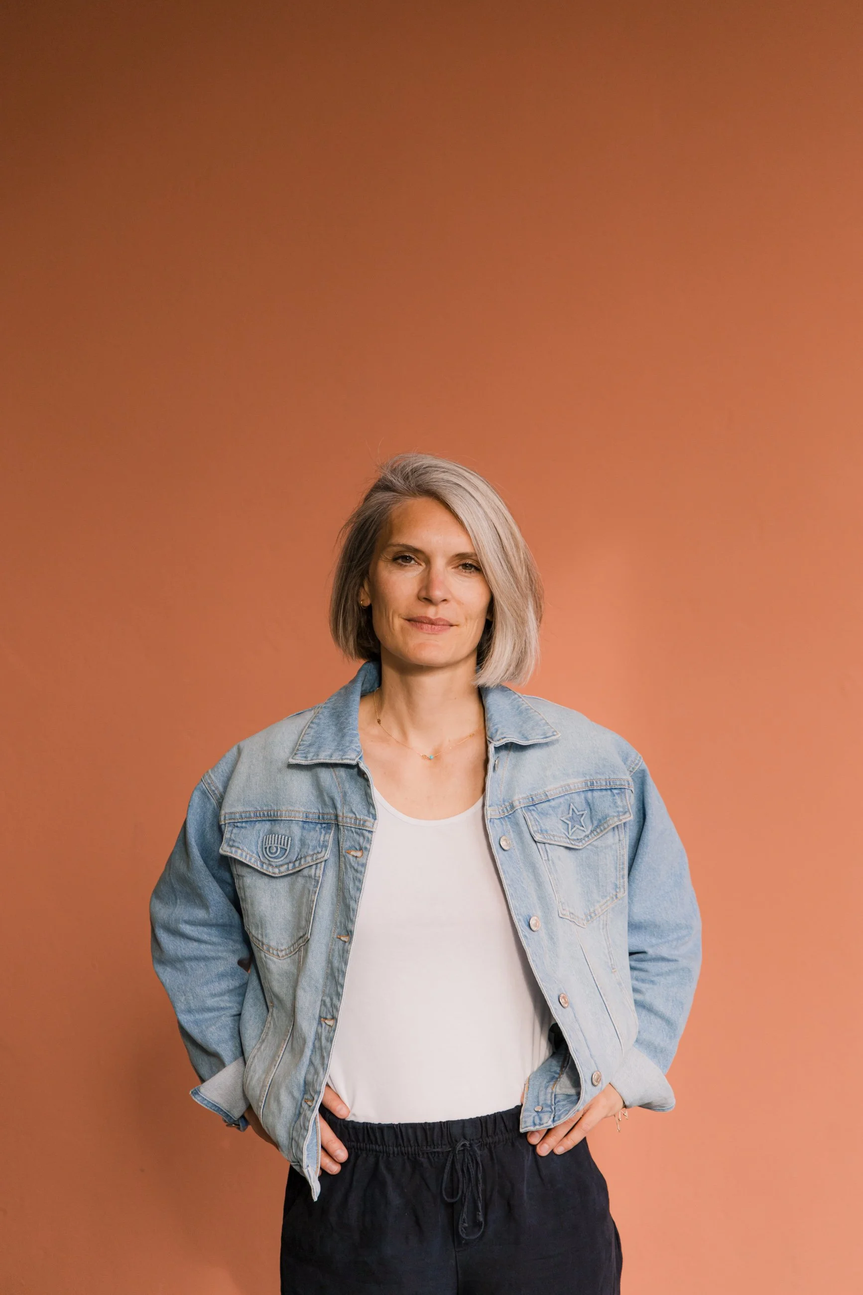 A woman with shoulder-length gray hair, wearing a denim jacket over a white t-shirt and dark pants, standing in front of a solid peach-colored background.