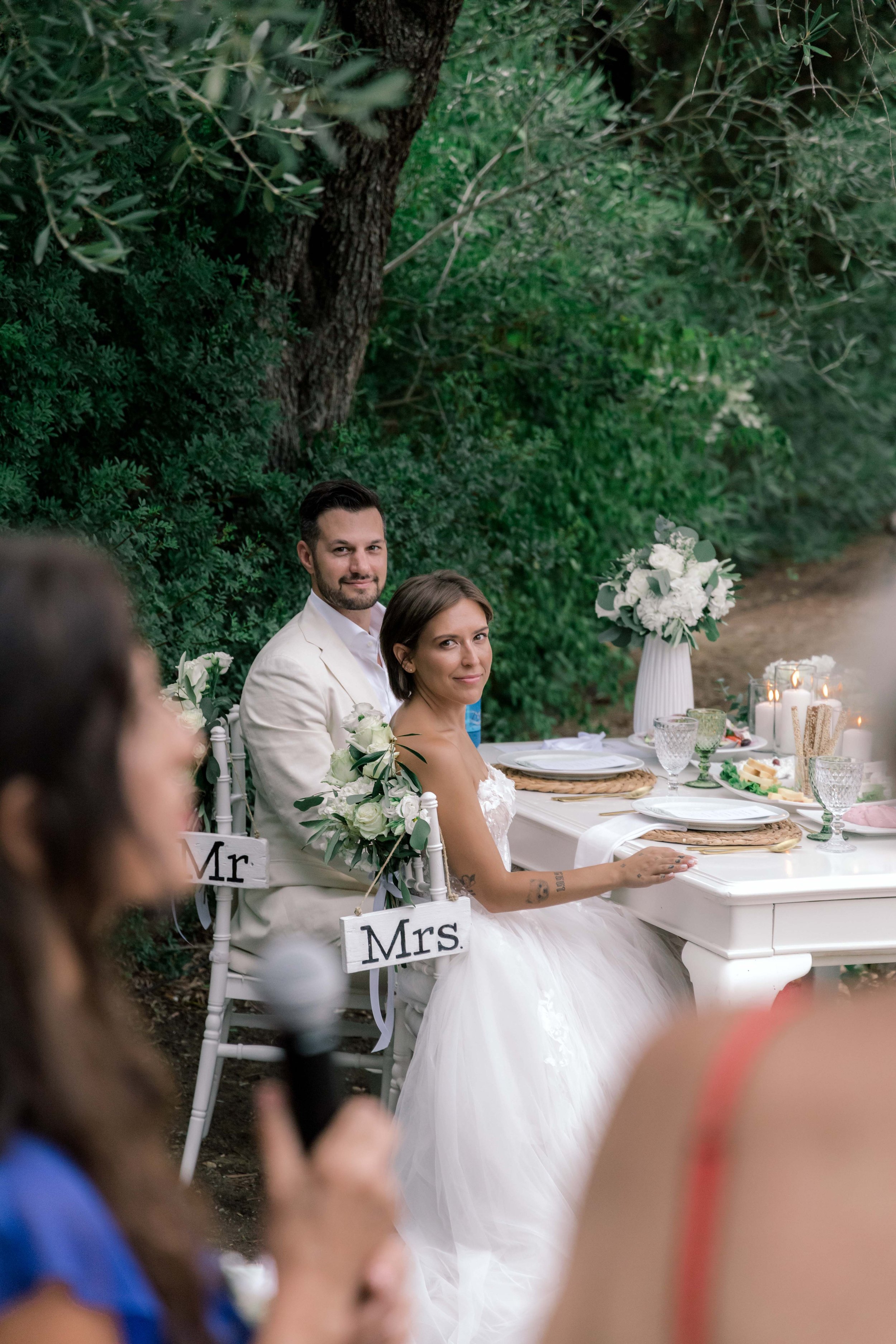 A newlywed couple, seated at a decorated outdoor wedding reception table, with the bride in a white wedding dress and the groom in a light-colored suit, surrounded by greenery.