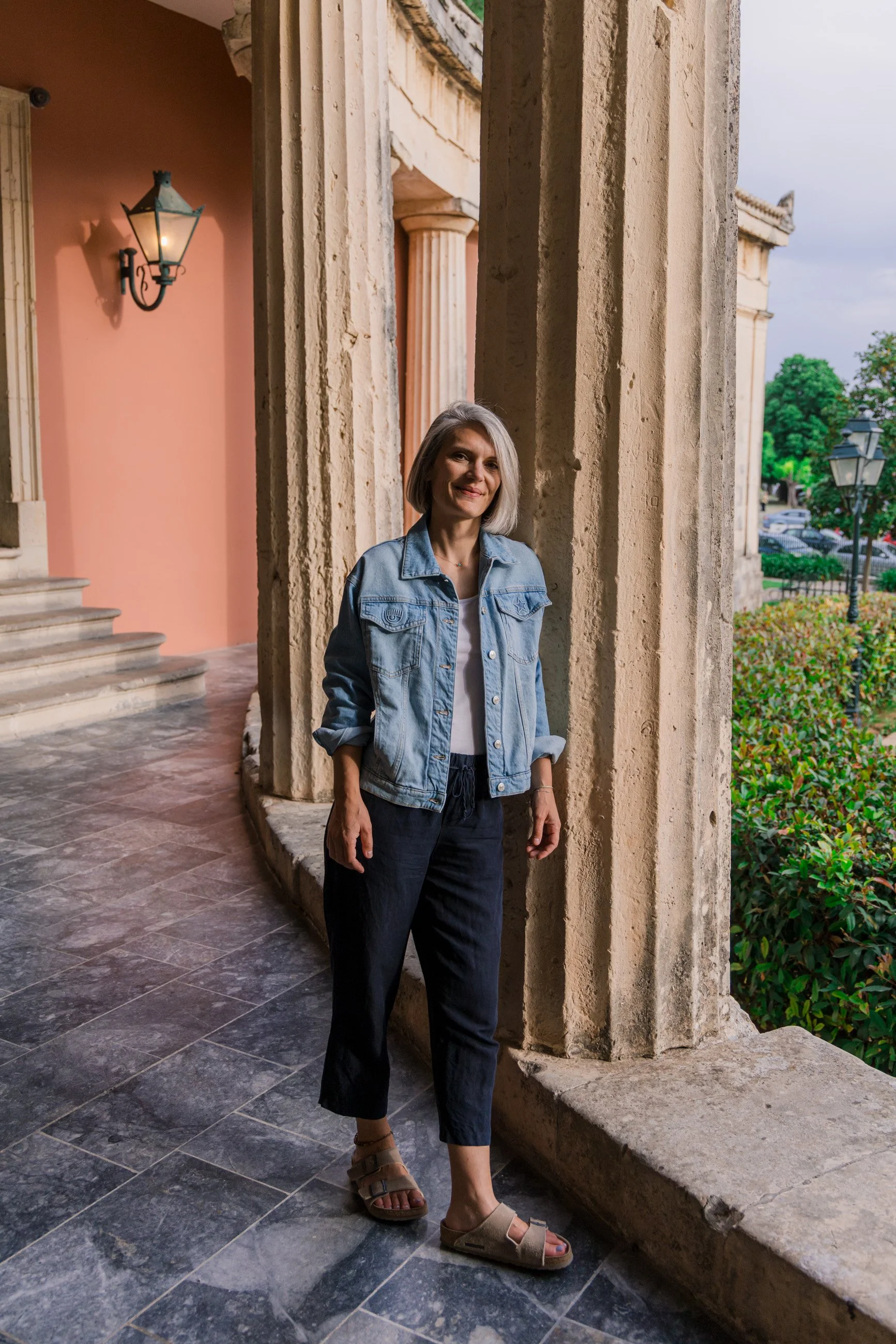 A woman standing on a stone-paved corridor near tall beige columns and a pink wall with a wall-mounted lamp, overlooking a green outdoor area with bushes and trees, during sunset.