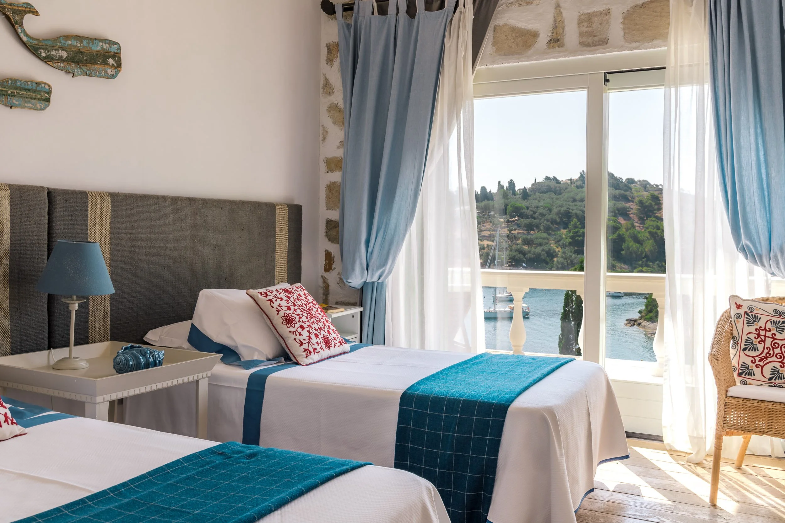 A bedroom with two beds, white linens, and blue accents. There is a bedside table with a blue lamp and decorative shell. A large window with sheer white curtains and blue drapes opens to a view of water, boats, and greenery outside.