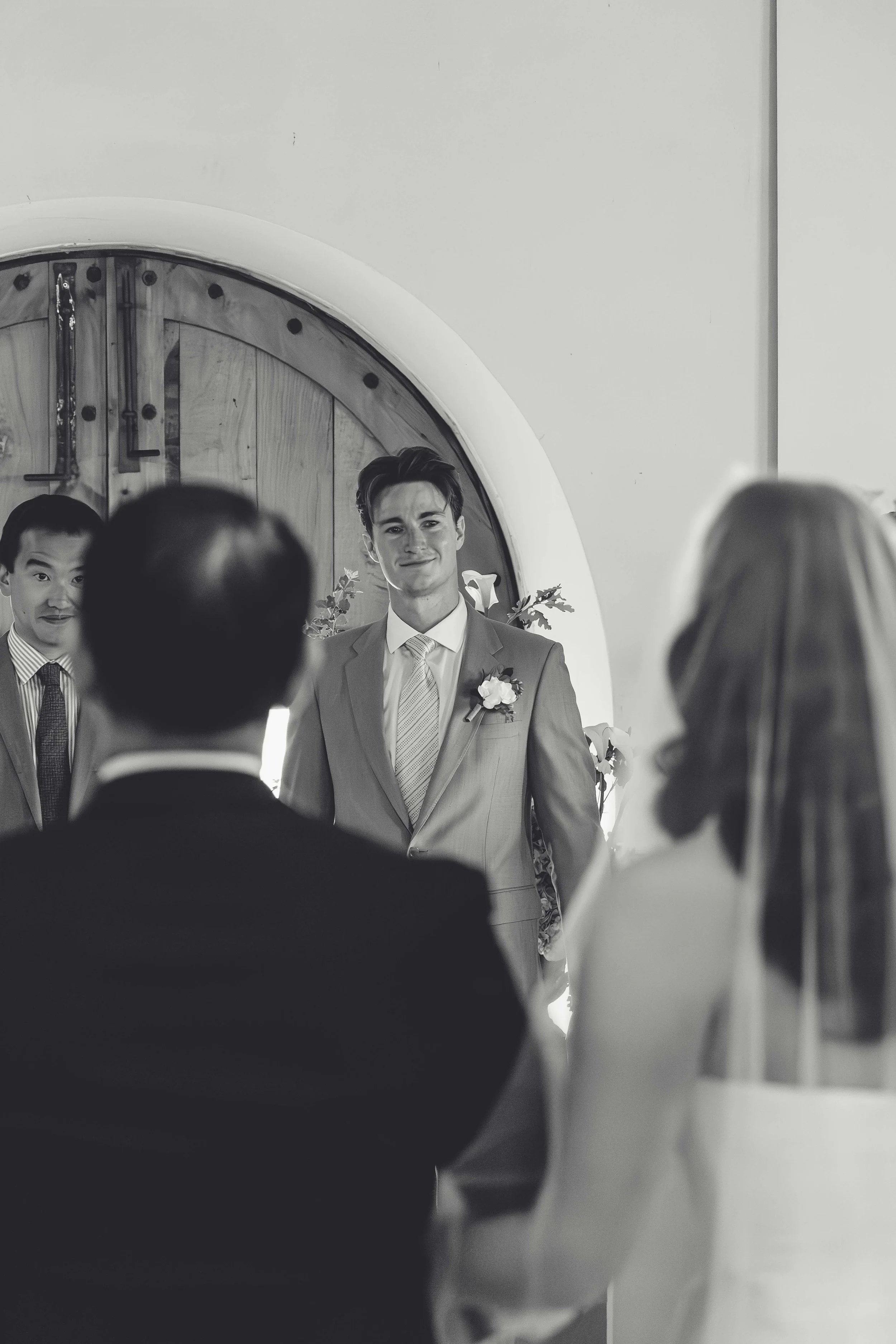 A black-and-white photo of a wedding ceremony showing a groom in a suit and tie standing in front of the altar, with his bride facing him, and two groomsmen standing to the side.