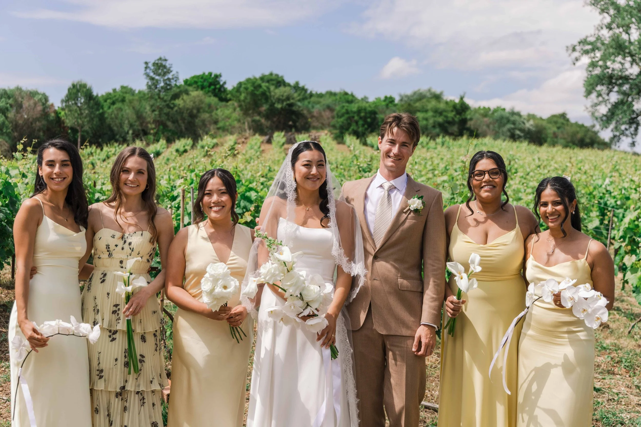 A wedding group photo of seven women and one man outdoors in a vineyard, with the bride in a white wedding dress and the groom in a beige suit, all smiling and holding white floral bouquets.