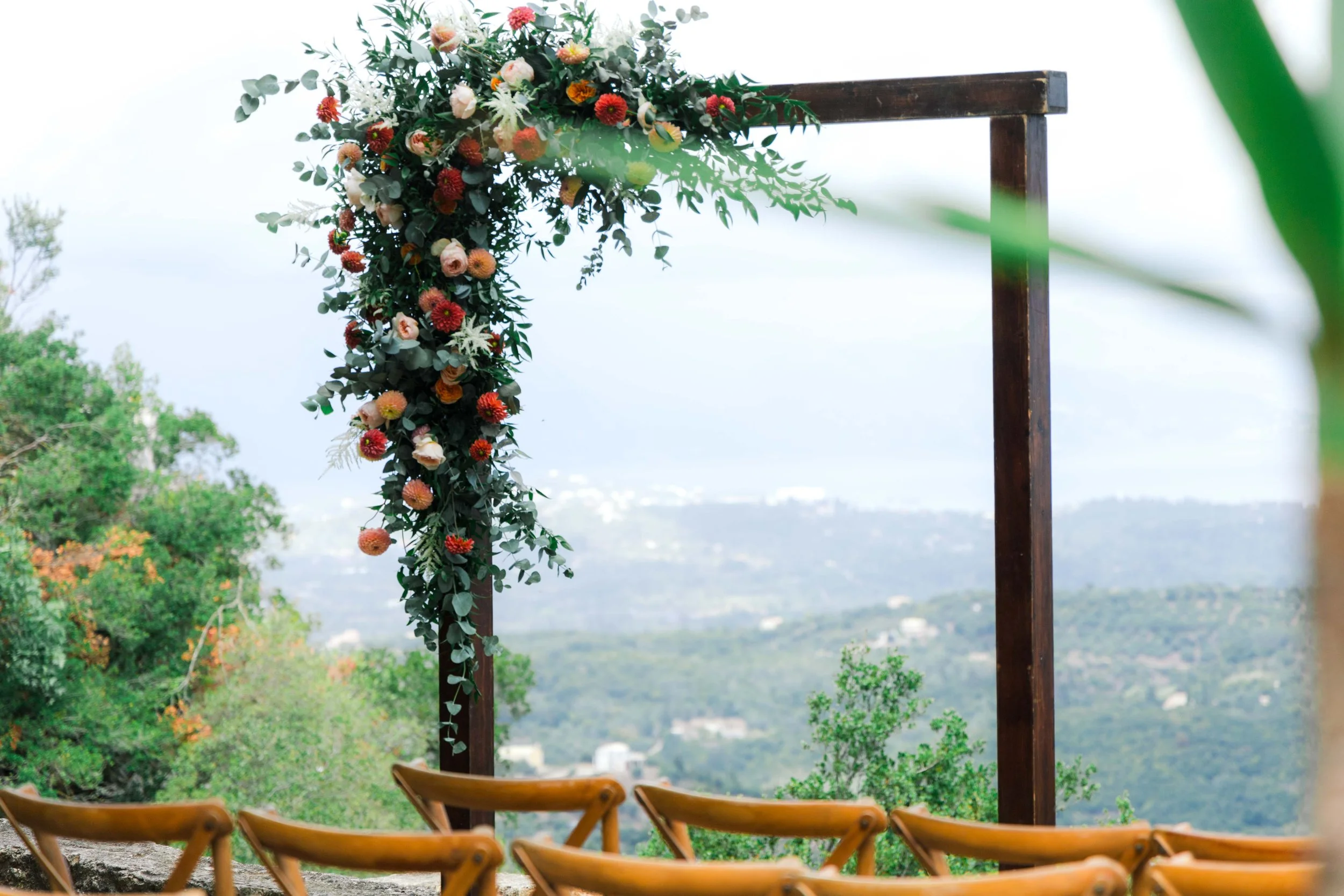 Wedding arch decorated with a floral arrangement of pink, red, and white flowers and greenery, set outdoors with mountain views and wooden chairs in the foreground.