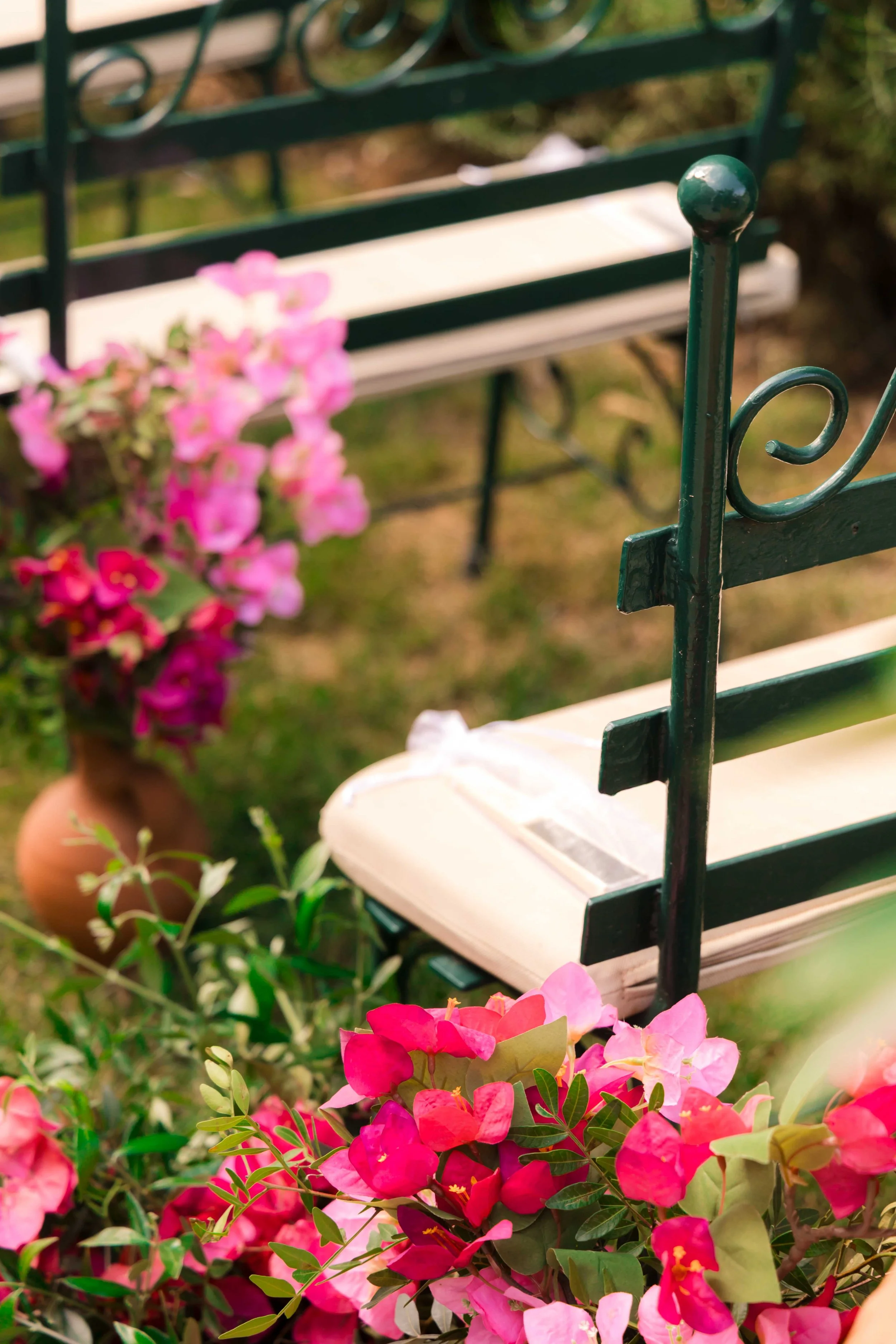 A white park bench with black metal armrests and legs, surrounded by pink and purple flowers in pots, in a garden setting.