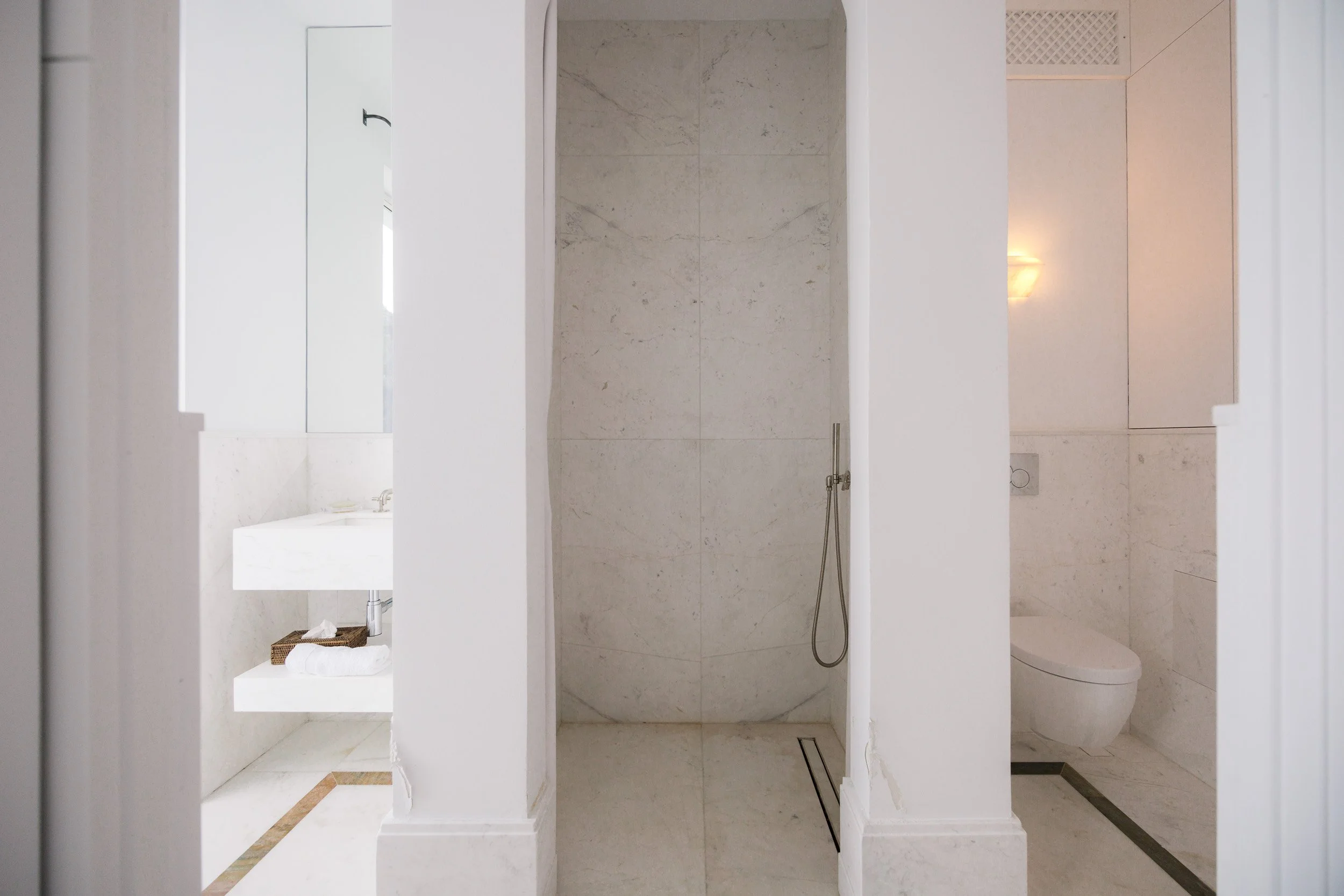View inside a modern bathroom with a shower in the center, a sink on the left with a basket of towels, and a toilet on the right, all in a light-colored marble finish.