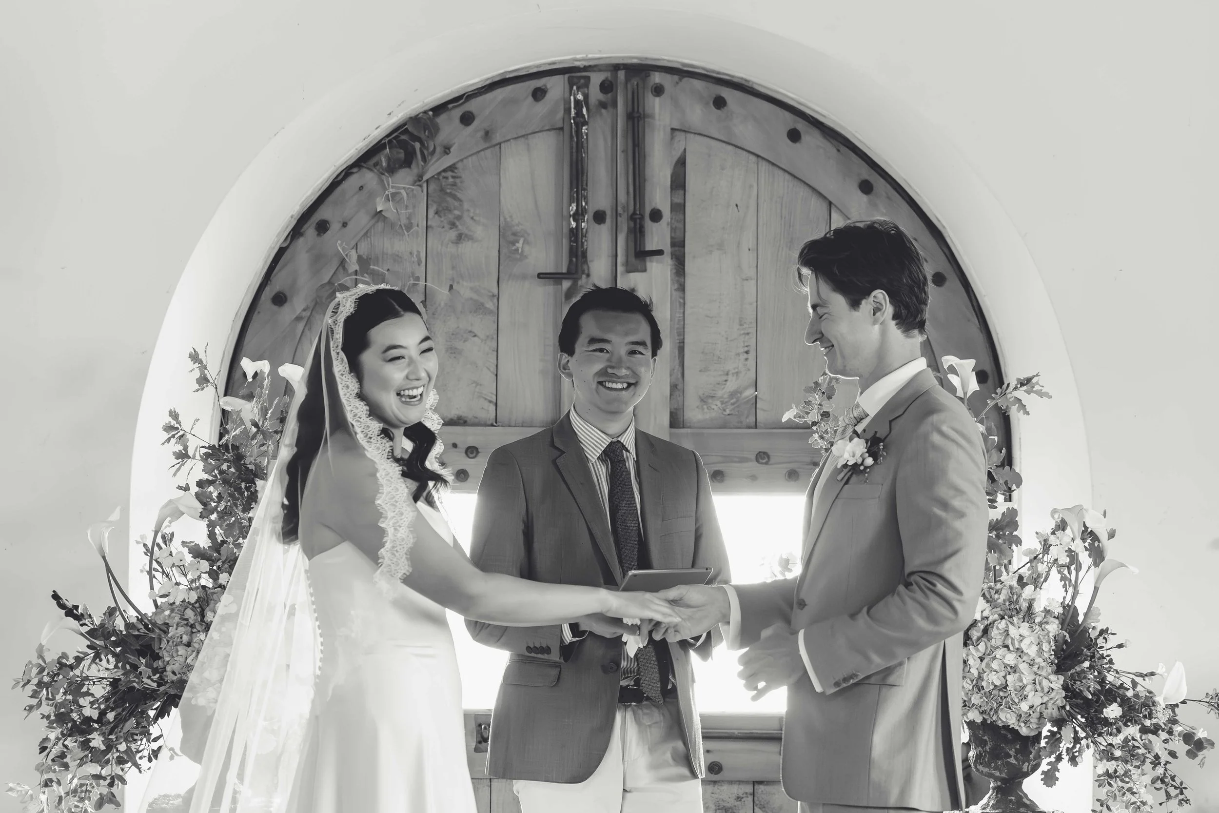 A wedding ceremony with a bride and groom holding hands, smiling at each other, and an officiant standing behind them in front of a wooden door with flowers on either side.