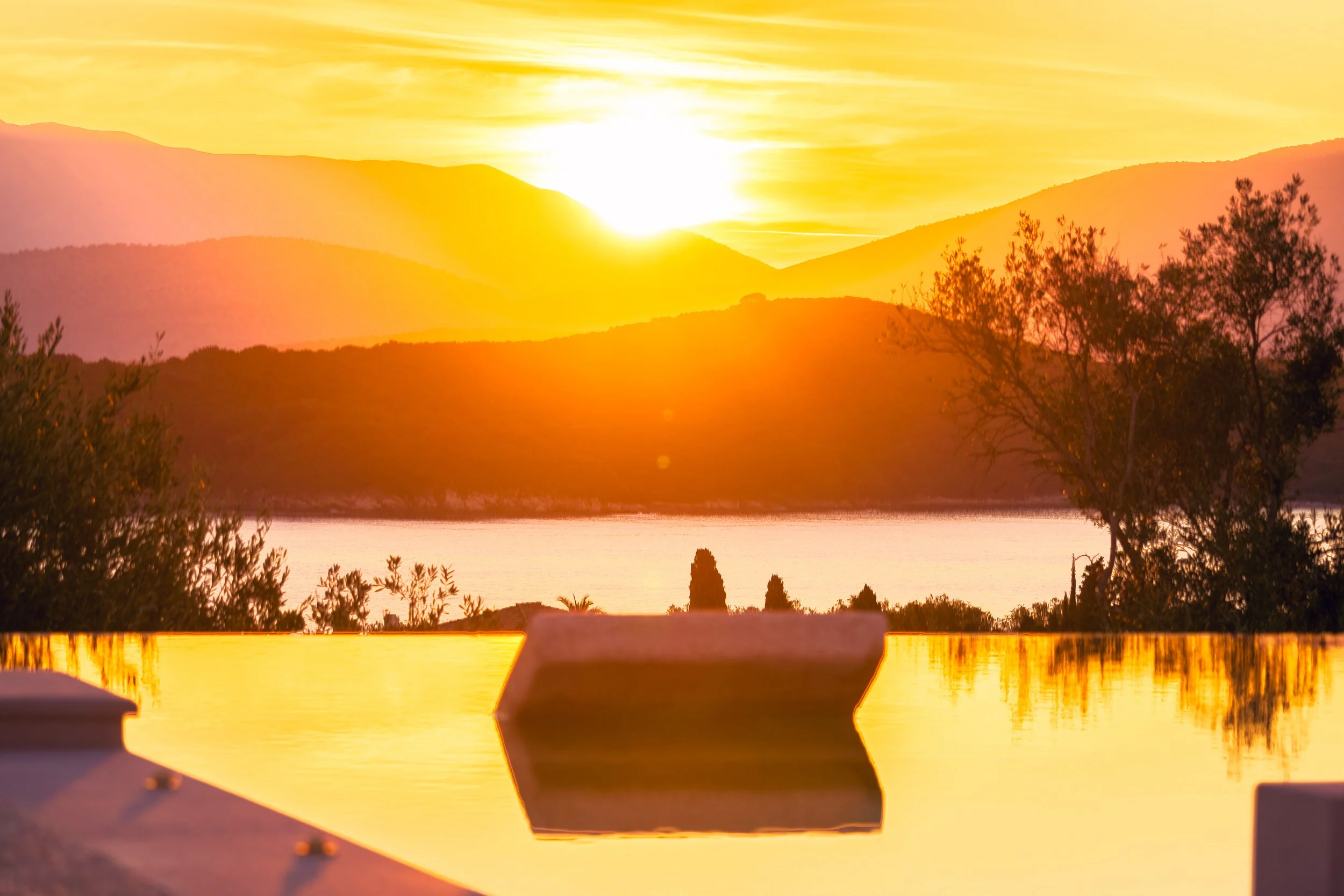 Sunset over mountains reflecting on a body of water with trees and a boat in the foreground.
