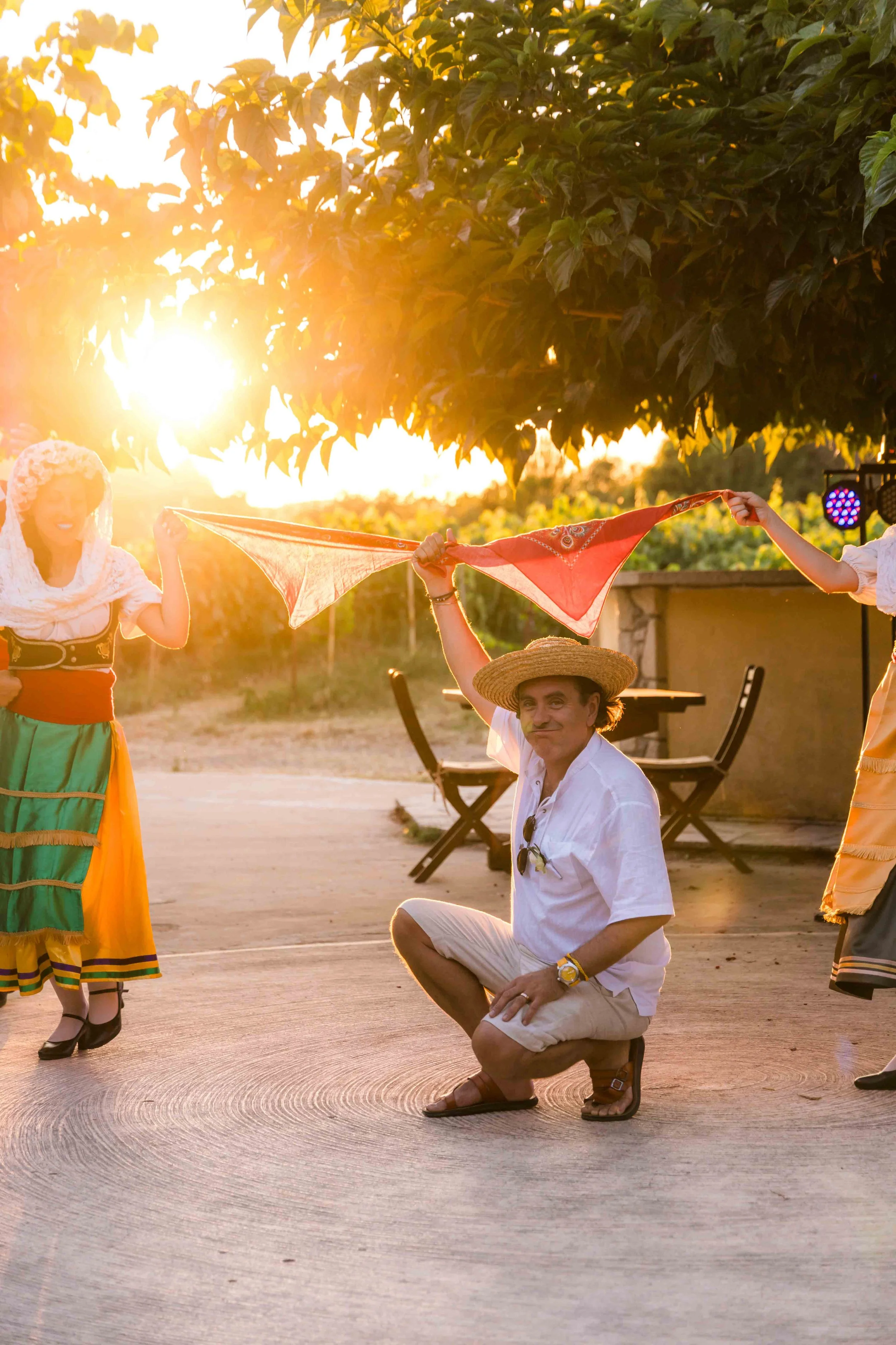People dancing outdoors during sunset, with one man crouching in the center wearing a straw hat and a white shirt, and women in colorful traditional dresses holding a cloth loop around him.