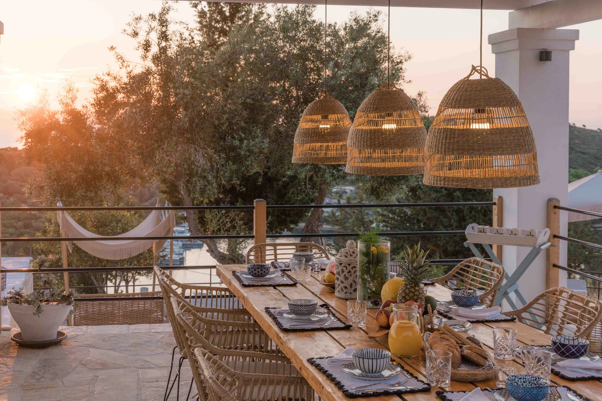 Outdoor dining table set for a meal at sunset with woven hanging pendant lights, surrounded by trees, with a hammock in the background.