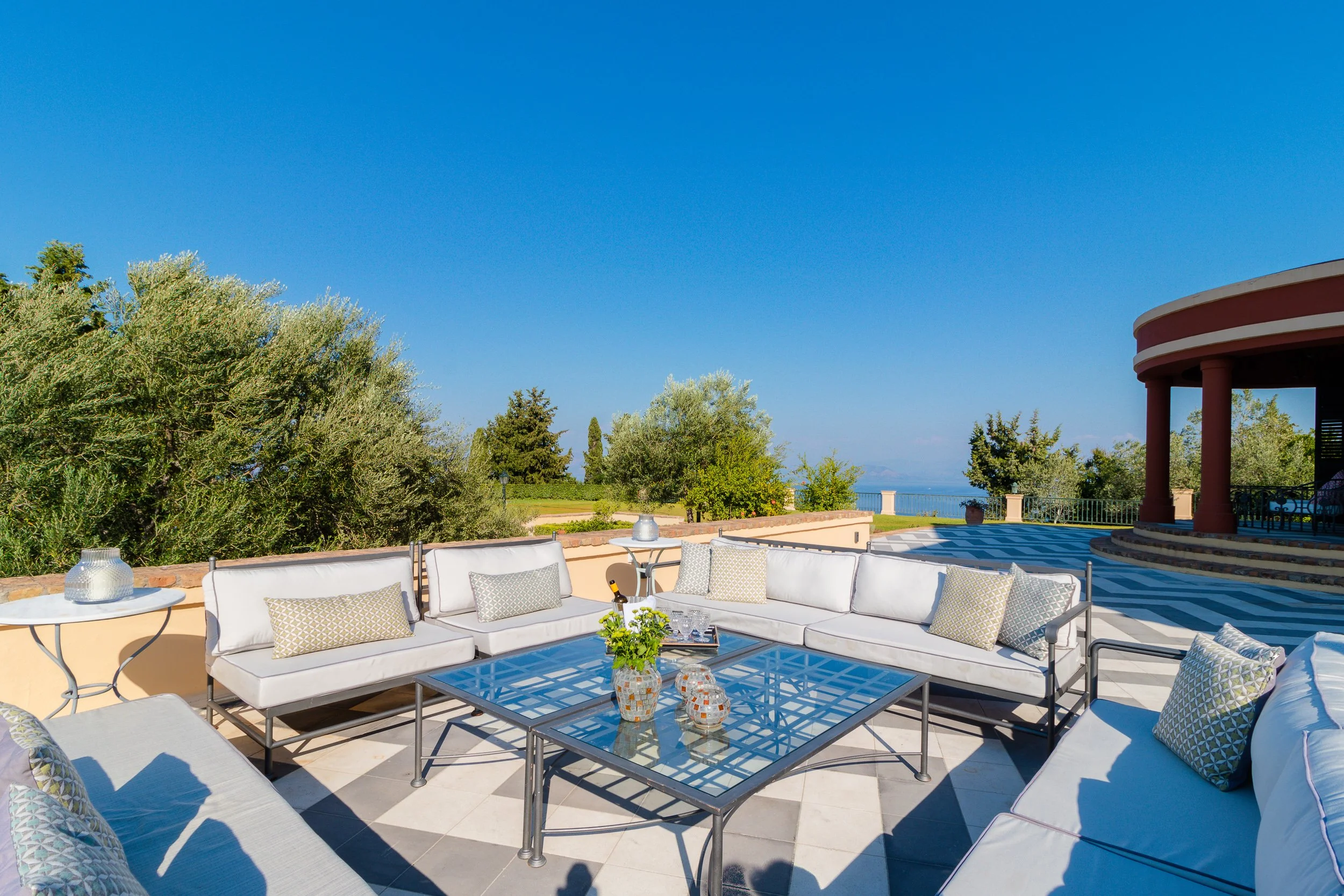 Outdoor patio with white sectional sofas, glass-topped coffee table, and decorative pillows under a bright blue sky, overlooking a lush garden and distant ocean view.