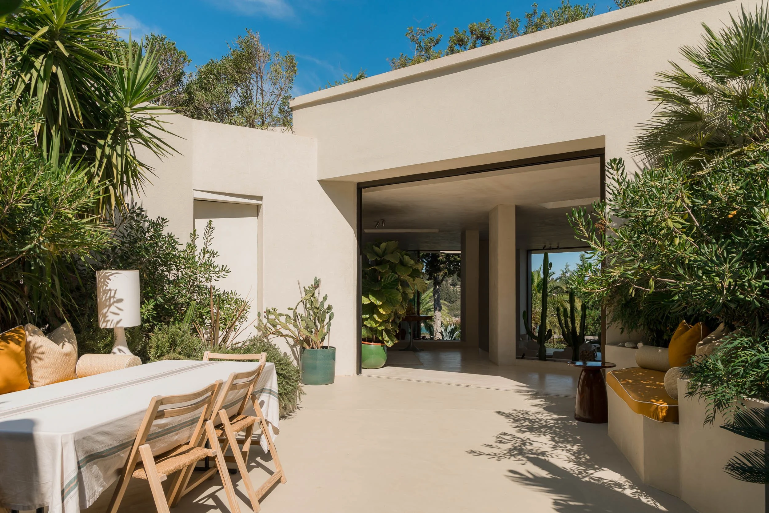Outdoor patio with white walls, greenery, a table with chairs, and cushioned benches with yellow pillows under a blue sky.