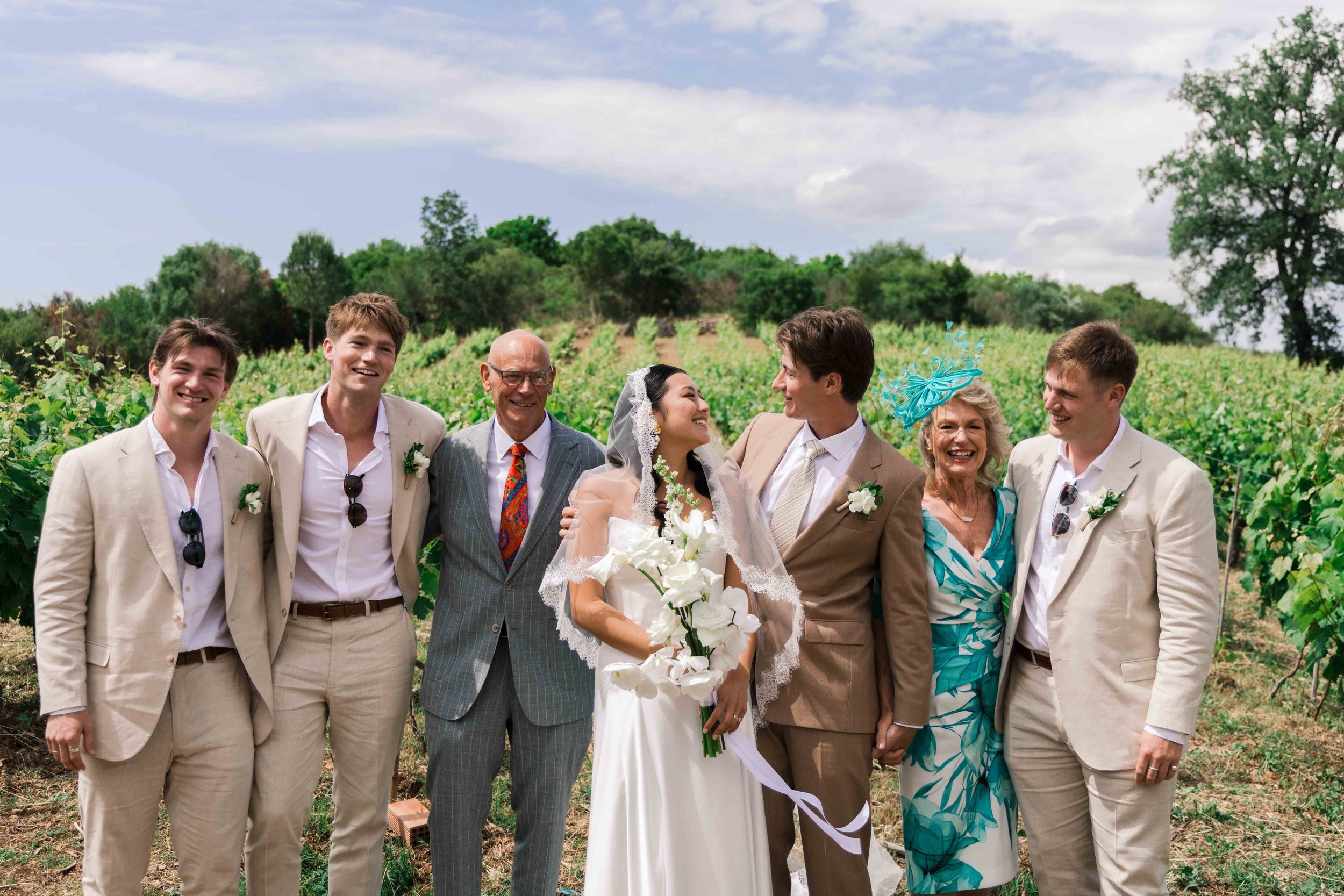 Group of wedding guests and a bride and groom smiling in a vineyard.
